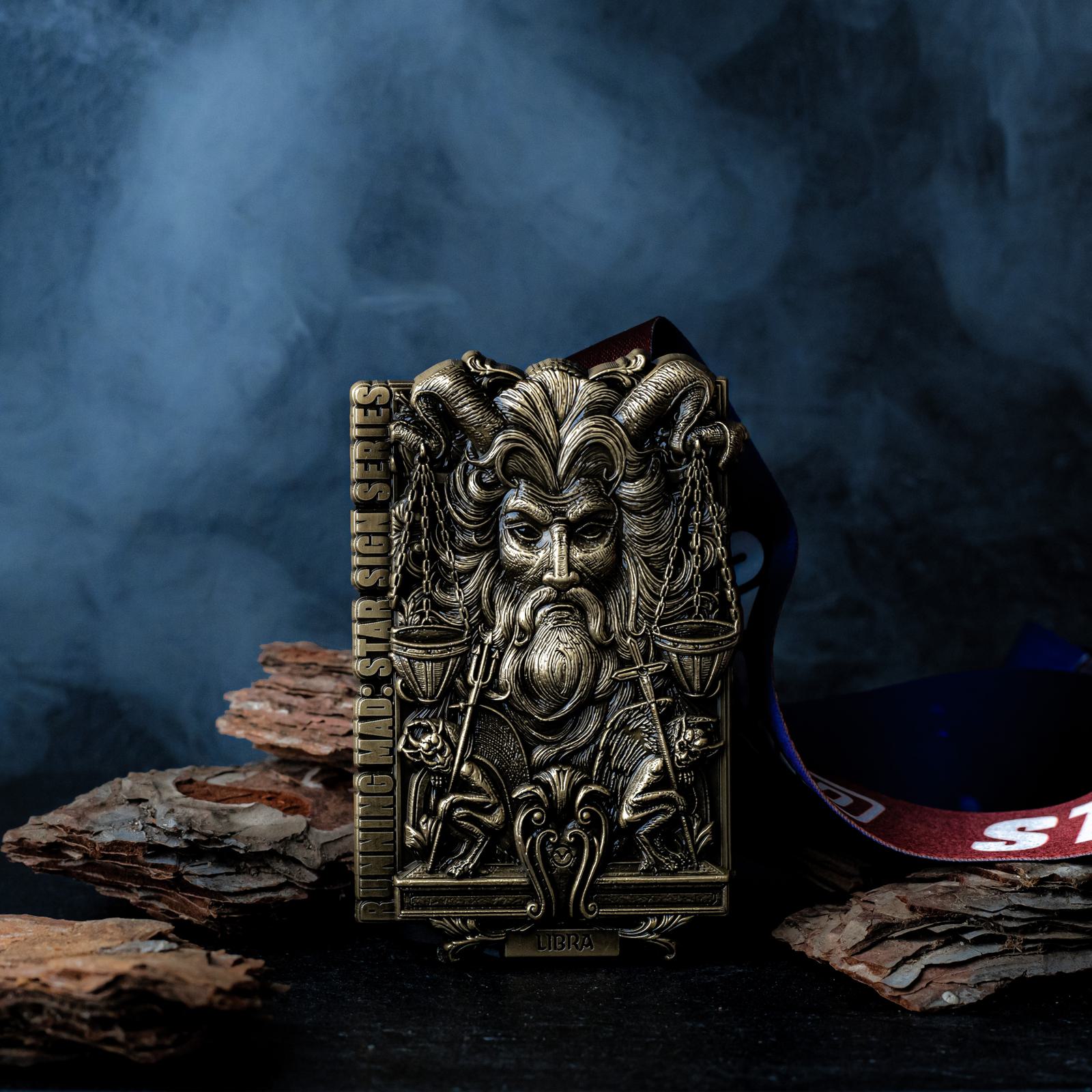 A detailed bronze medallion depicts a mythical figure with horns, a beard, and a scale. The background is smoky, with pieces of bark on a dark surface. A ribbon extends from the medallion, partially visible with text.