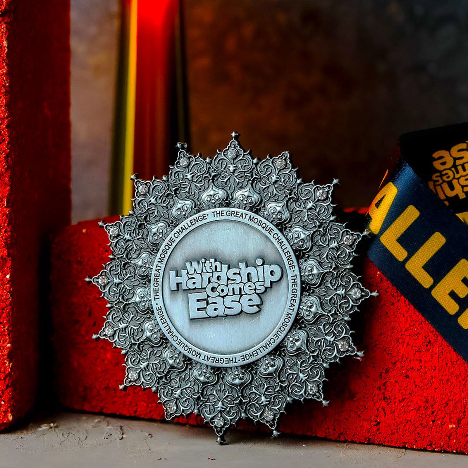 A silver medal with intricate floral designs features the inscription "With Hardship Comes Ease" in the center. The medal is displayed against a red textured background.