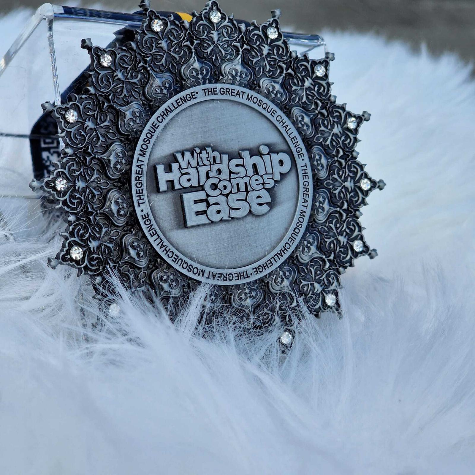 A detailed circular medal with an ornate border rests on a white, fluffy surface. The medal features the text "With Hardship Comes Ease" and "The Great Mosque Challenge" repeated around the edge.
