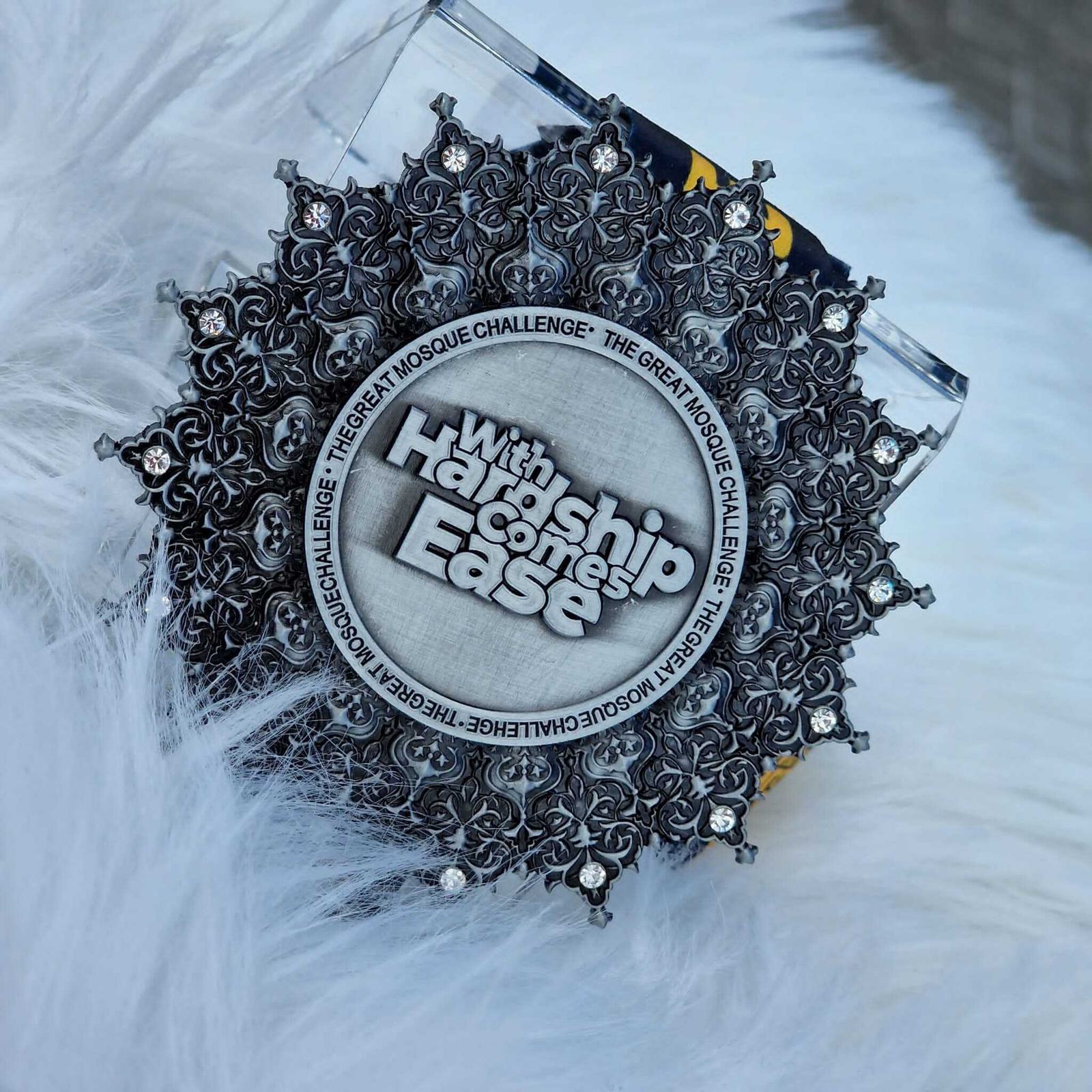 An intricately designed silver medallion placed on a soft white fur surface. The medallion is inscribed with the words "With Hardship Comes Ease." It is surrounded by ornate patterns and gemstones.