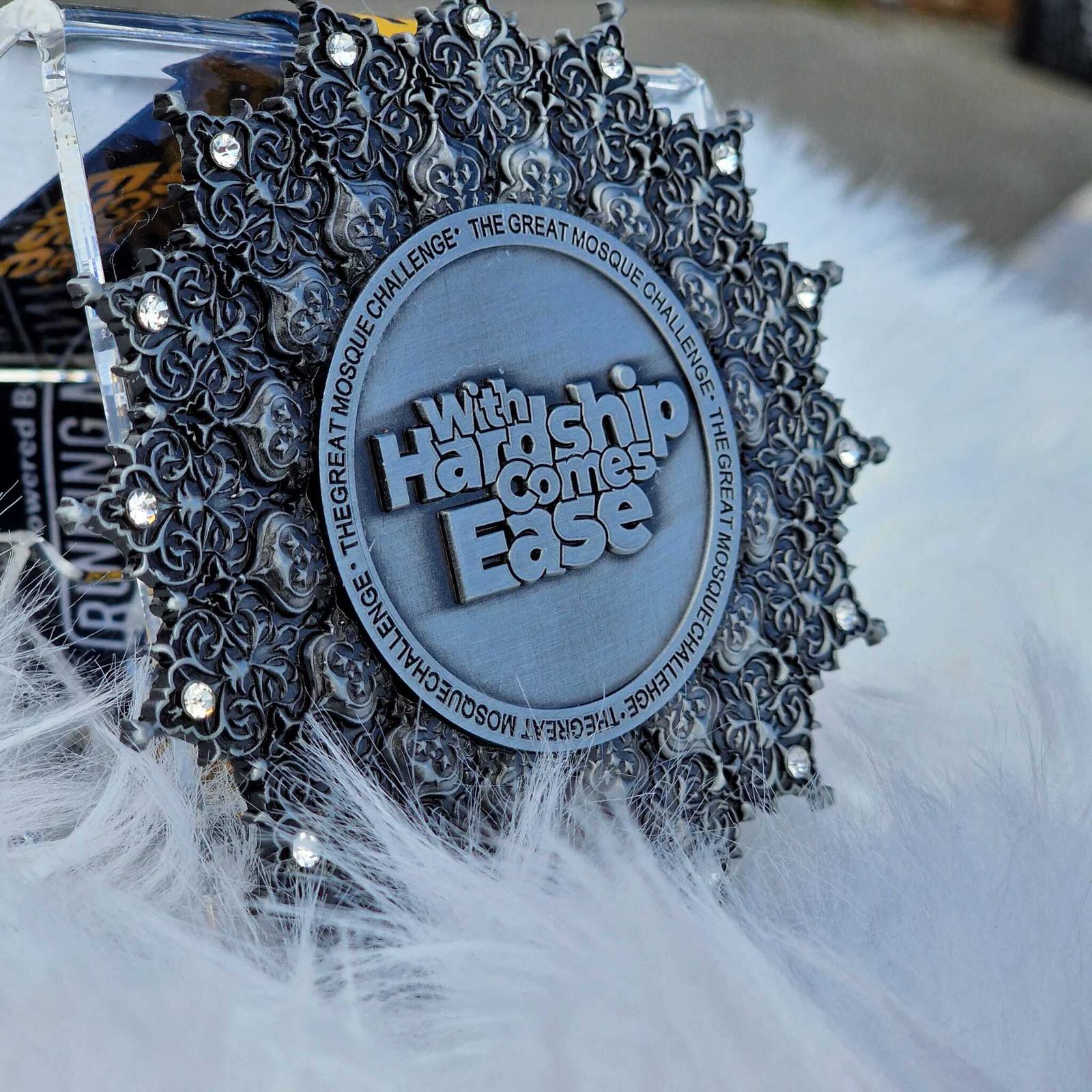 A decorative medal with intricate silver patterns surrounds the words "With Hardship Comes Ease." The border contains text referring to "The Great Mosque Challenge." The medal is placed on a soft, fluffy white surface.