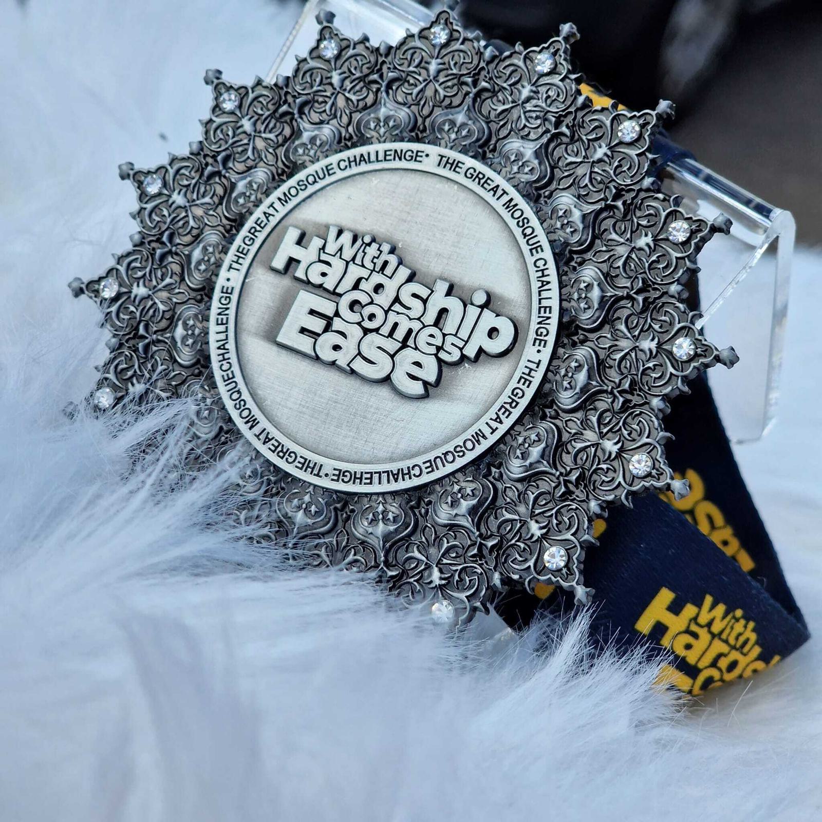 A beautifully ornate round medal with the inscription "With Hardship Comes Ease" is displayed on a soft, fluffy white surface. The medal features intricate detailing and a ribbon with the phrase "The Great Mosque Challenge.
