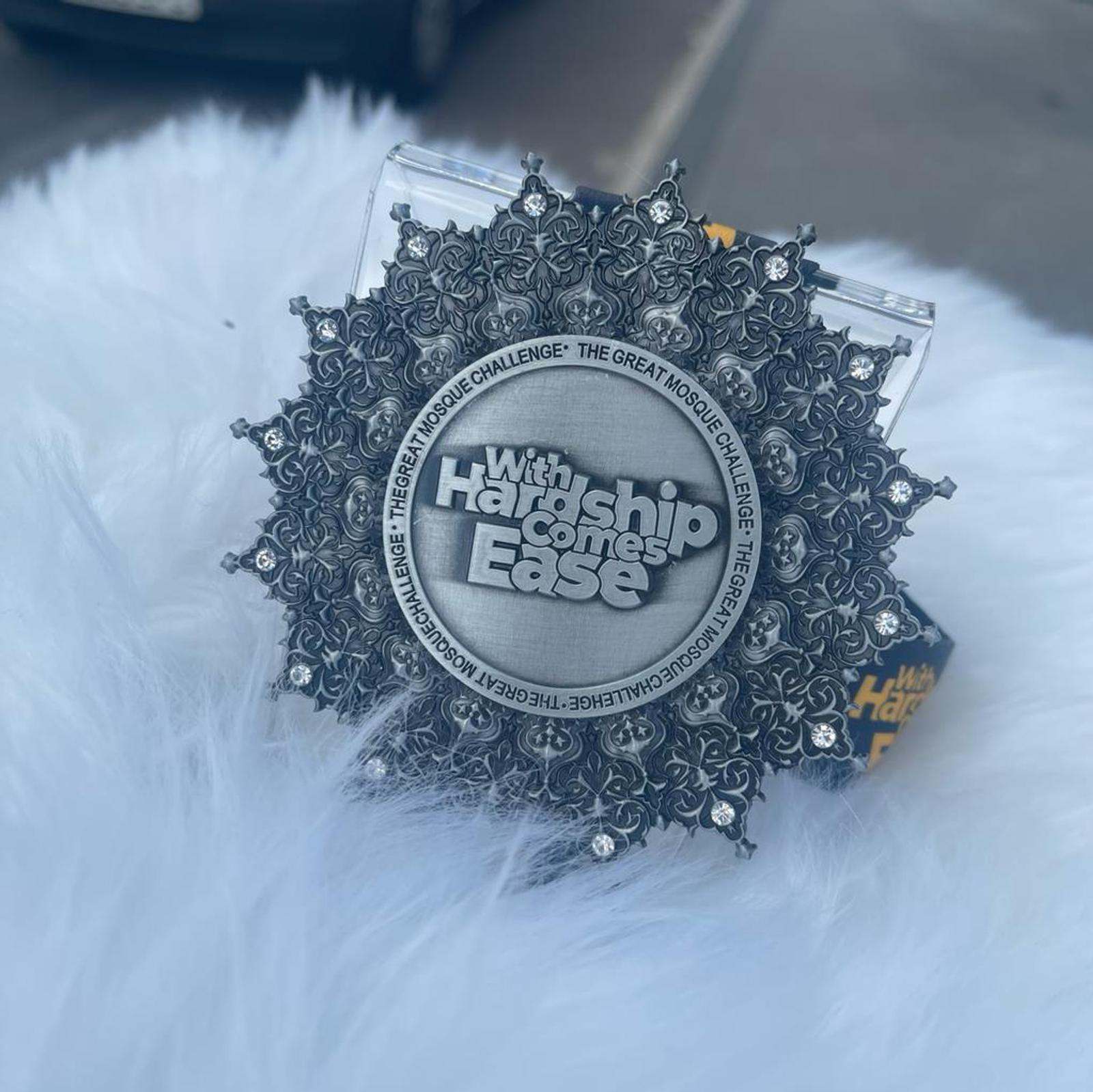 A decorative silver medal with intricate floral designs and the text "With Hardship Comes Ease" in the center. The edge reads, "The Great Mosque Challenge." The medal rests on a fluffy white surface.