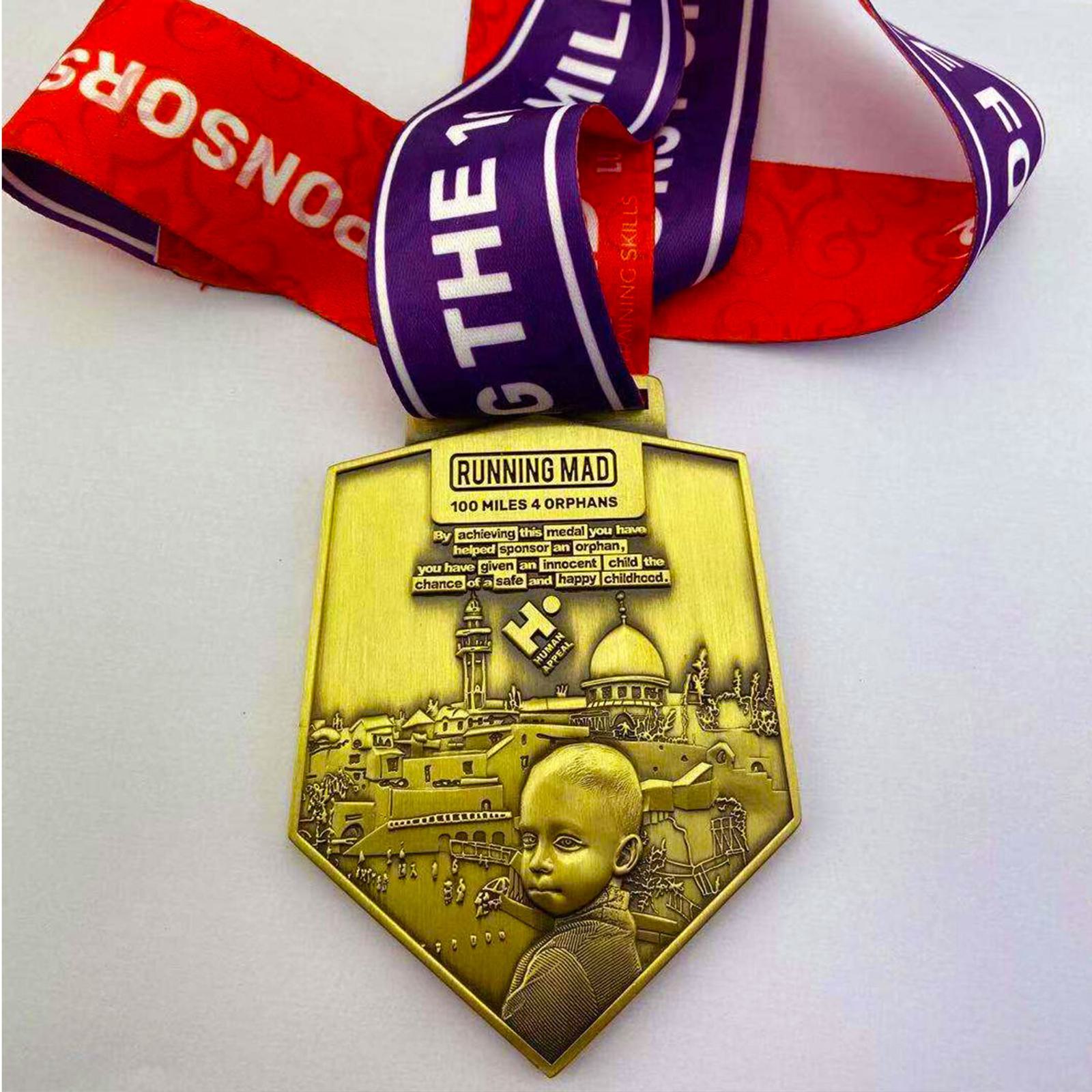 A gold medal with an embossed design featuring a child and cityscape. The medal reads "Running Mad 100 Miles 4 Orphans." It hangs from a purple and red ribbon printed with white text.