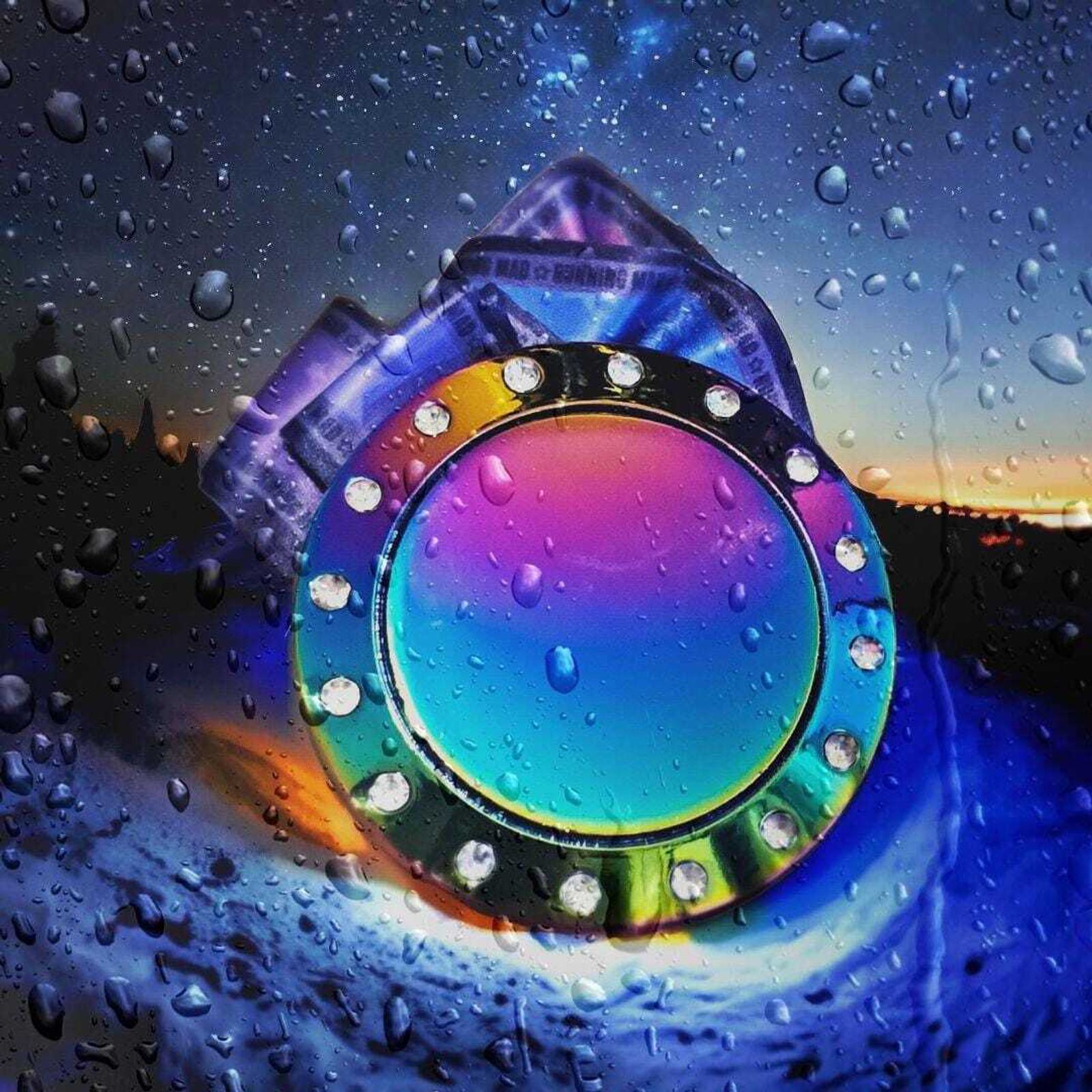 A colorful, circular object with a reflective surface and small stones around its border is set against a dark, starry sky. Water droplets on the surface create a dramatic, dreamy effect.