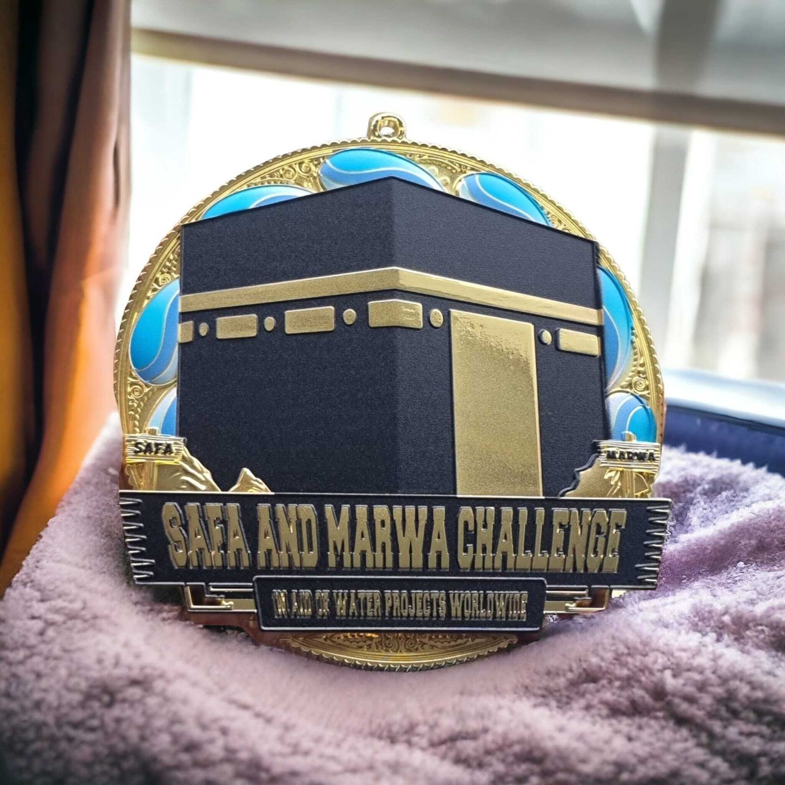 A medal featuring a design resembling the Kaaba, with blue accents and the text "Safa and Marwa Challenge" and "In Aid of Water Projects Worldwide." It rests on a soft pink surface near a window.