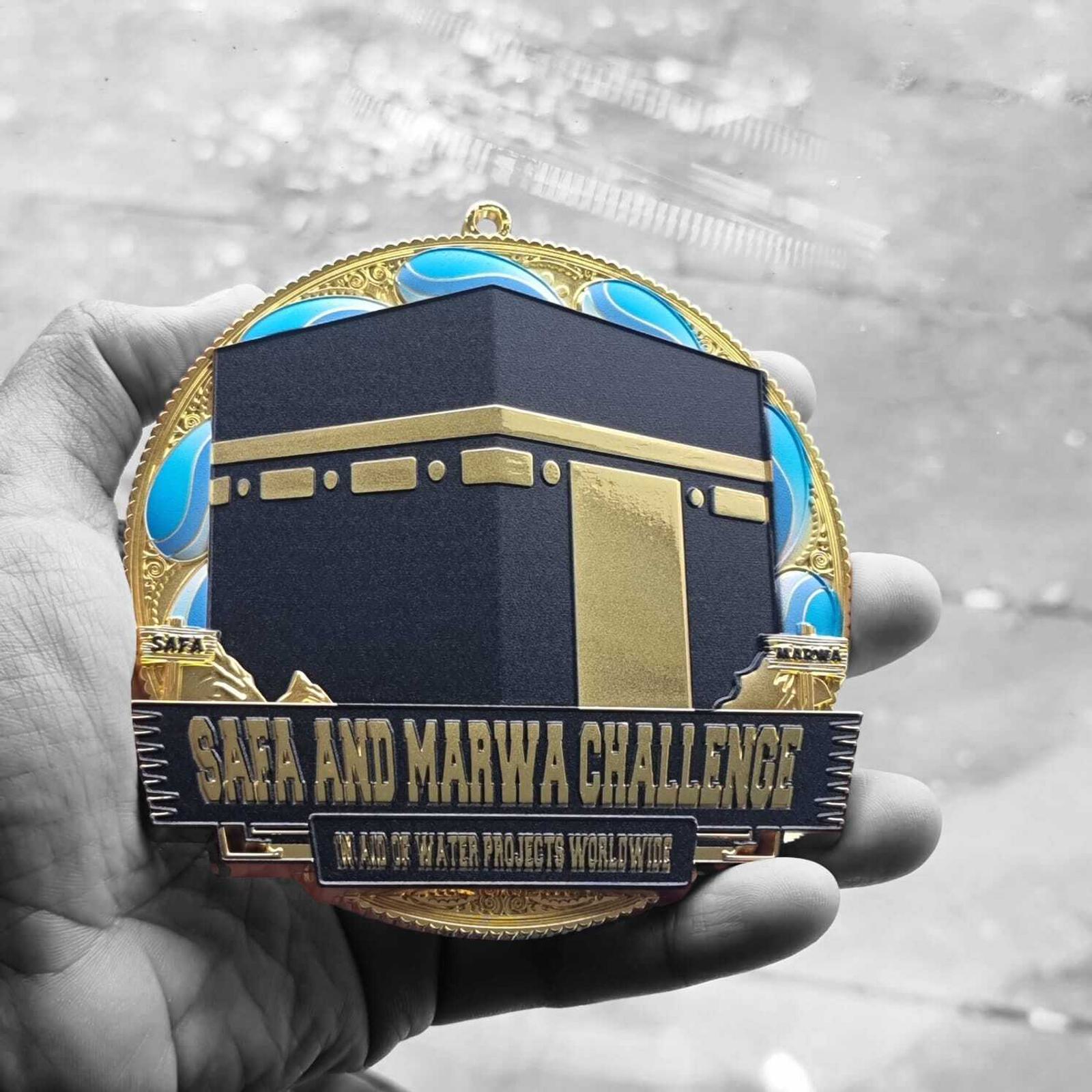 A hand holds a medal featuring a gold and black illustration of the Kaaba, surrounded by a decorative border with blue accents. The text reads "Safa and Marwa Challenge" and "To Aid in Water Projects Worldwide.