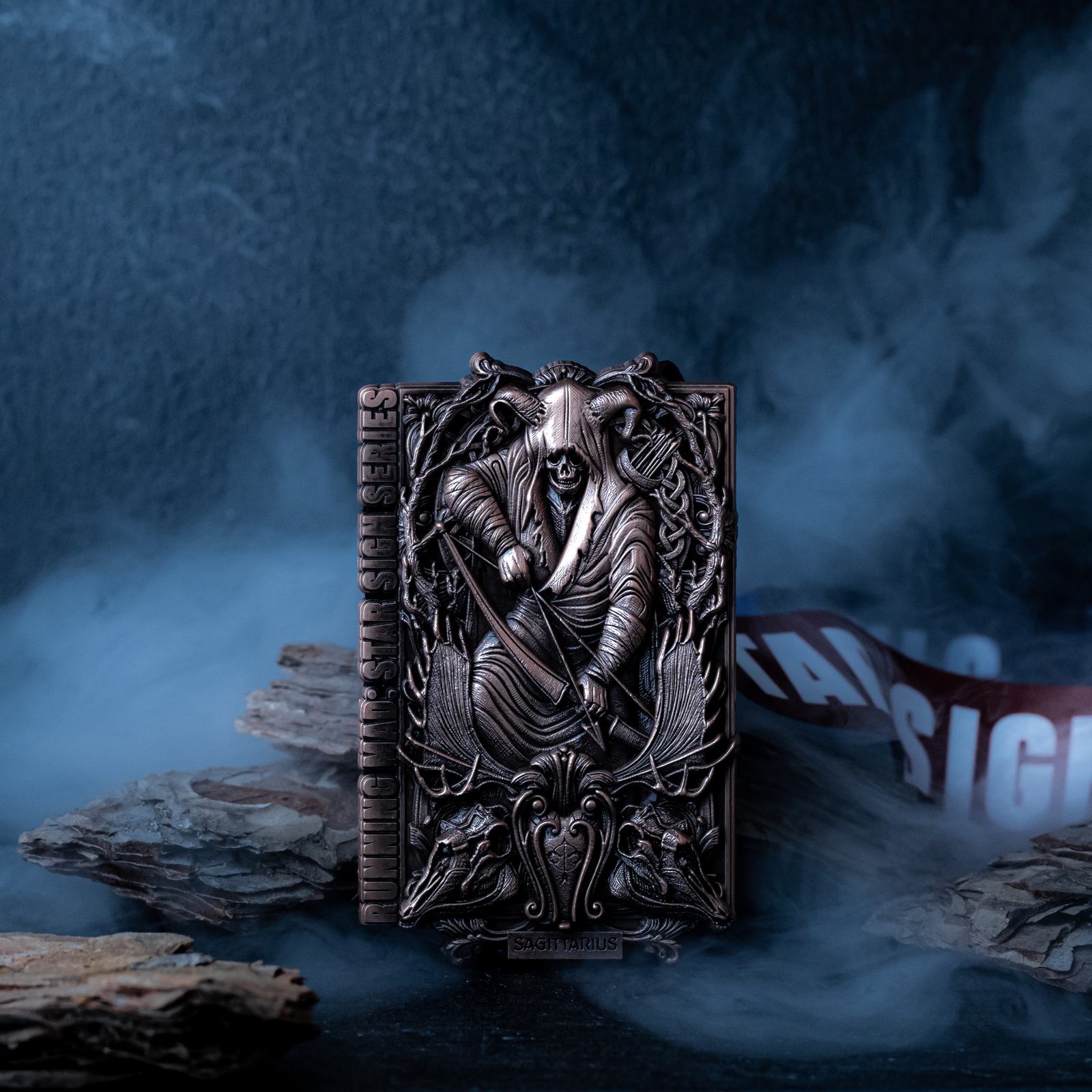 A detailed, embossed metal card features a robed figure holding a sword, surrounded by intricate designs. The card is labeled "Sagittarius" and is placed on a misty surface with bark pieces.
