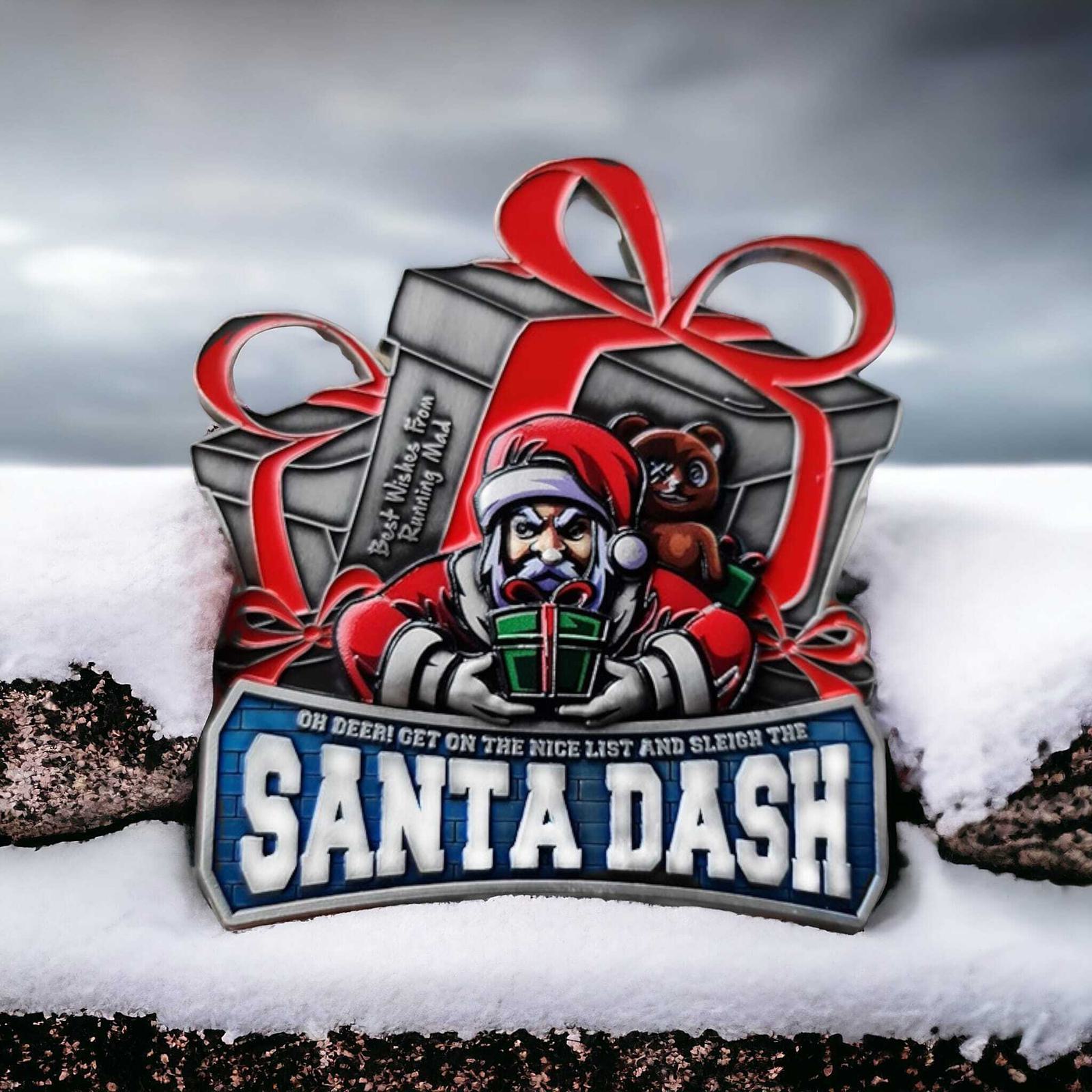 A Santa-themed medal rests on snow, featuring Santa holding gifts, a teddy bear peeking from a wrapped present, and the text "Santa Dash" in bold blue letters. The sky is overcast in the background.