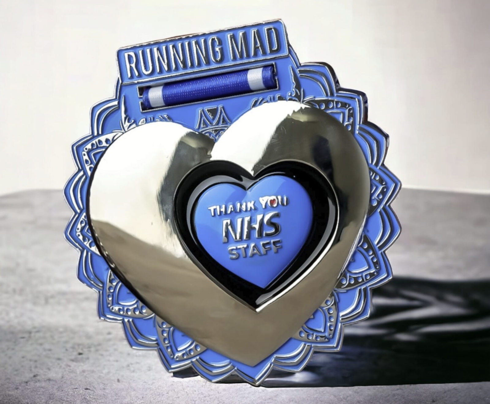A decorative medal with a metallic heart centerpiece and a bold "Thank You NHS Staff" inscription. Adorned with intricate blue patterns, the top of the medal reads "Running Mad." The medal embodies appreciation for healthcare workers.