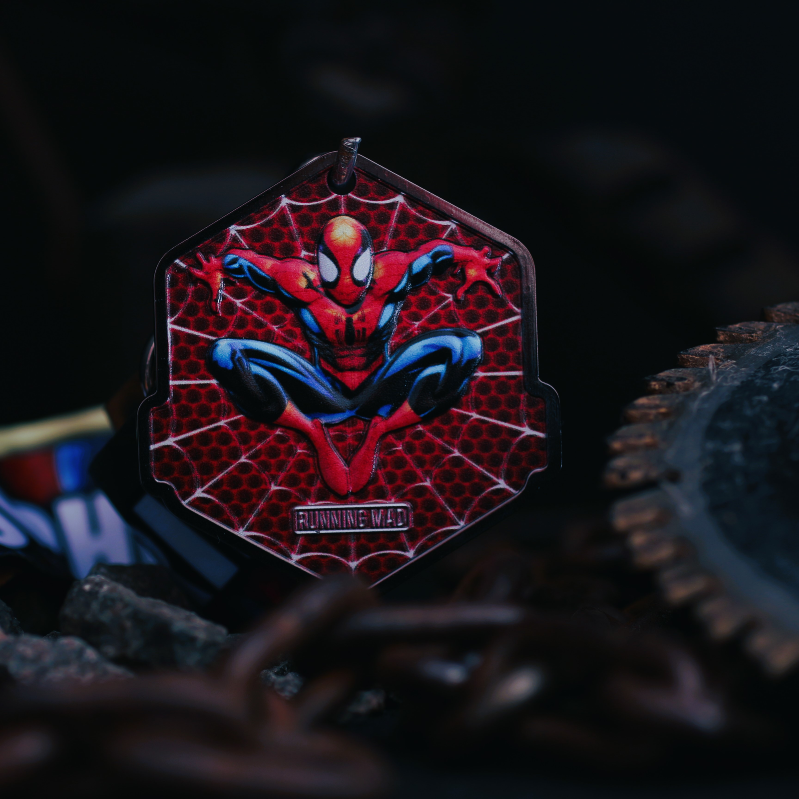 A keychain featuring a colorful illustration of a superhero in a red and blue suit, set against a web-patterned background. It hangs among chains and gears in a dark setting.