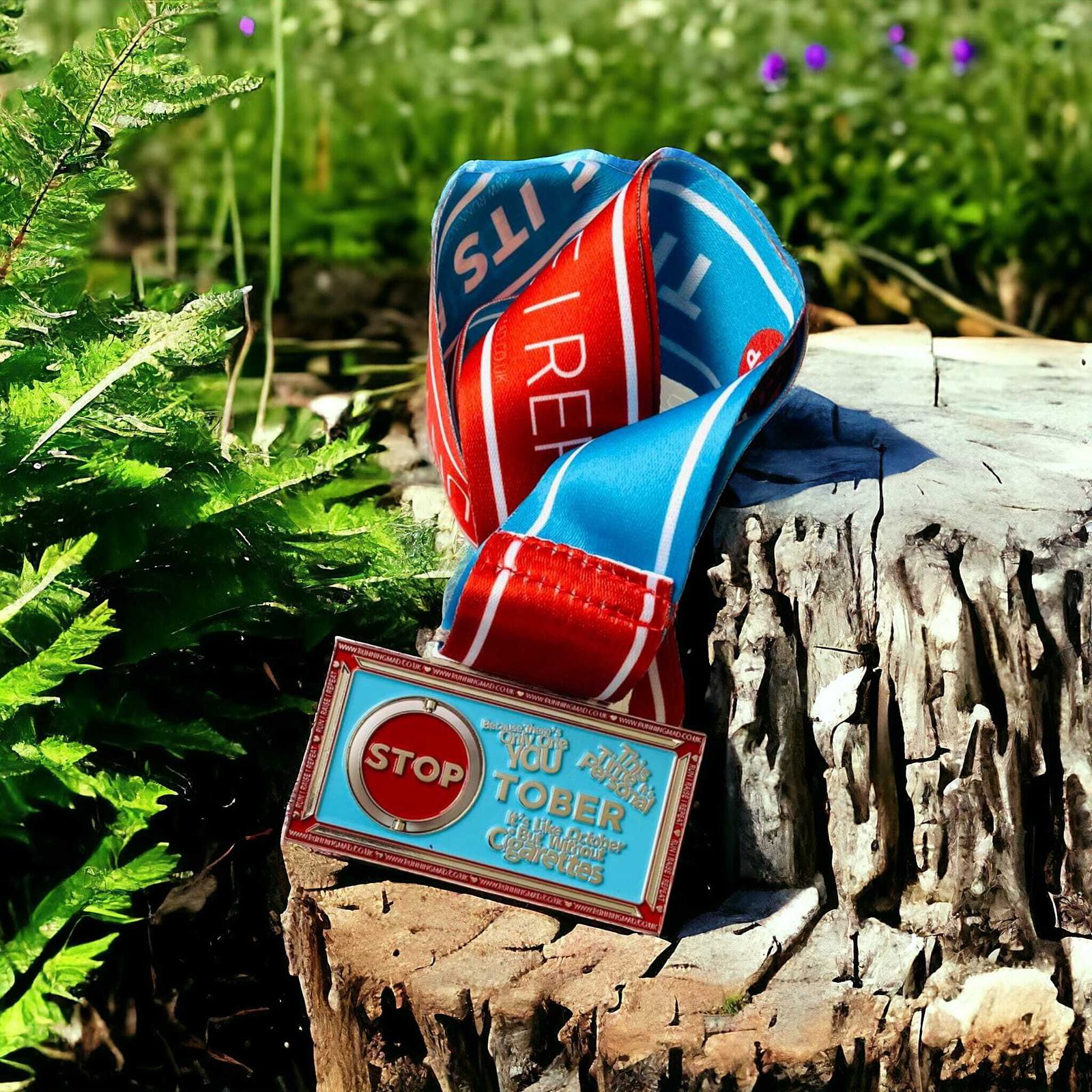 A colorful medal with "STOP" in bold letters and "TOBER" below is draped over a tree stump. The ribbon is blue and red with white text. The backdrop features greenery and purple flowers under bright sunlight.