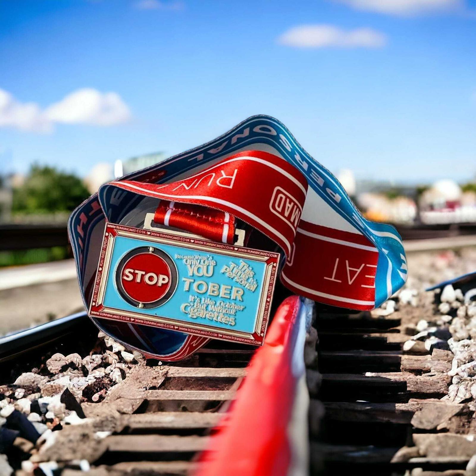 A "STOP" sign belt lies on a railway track against a bright blue sky. The belt is adorned with motivational text and colorful patterns, partially covering the wooden and metal tracks.