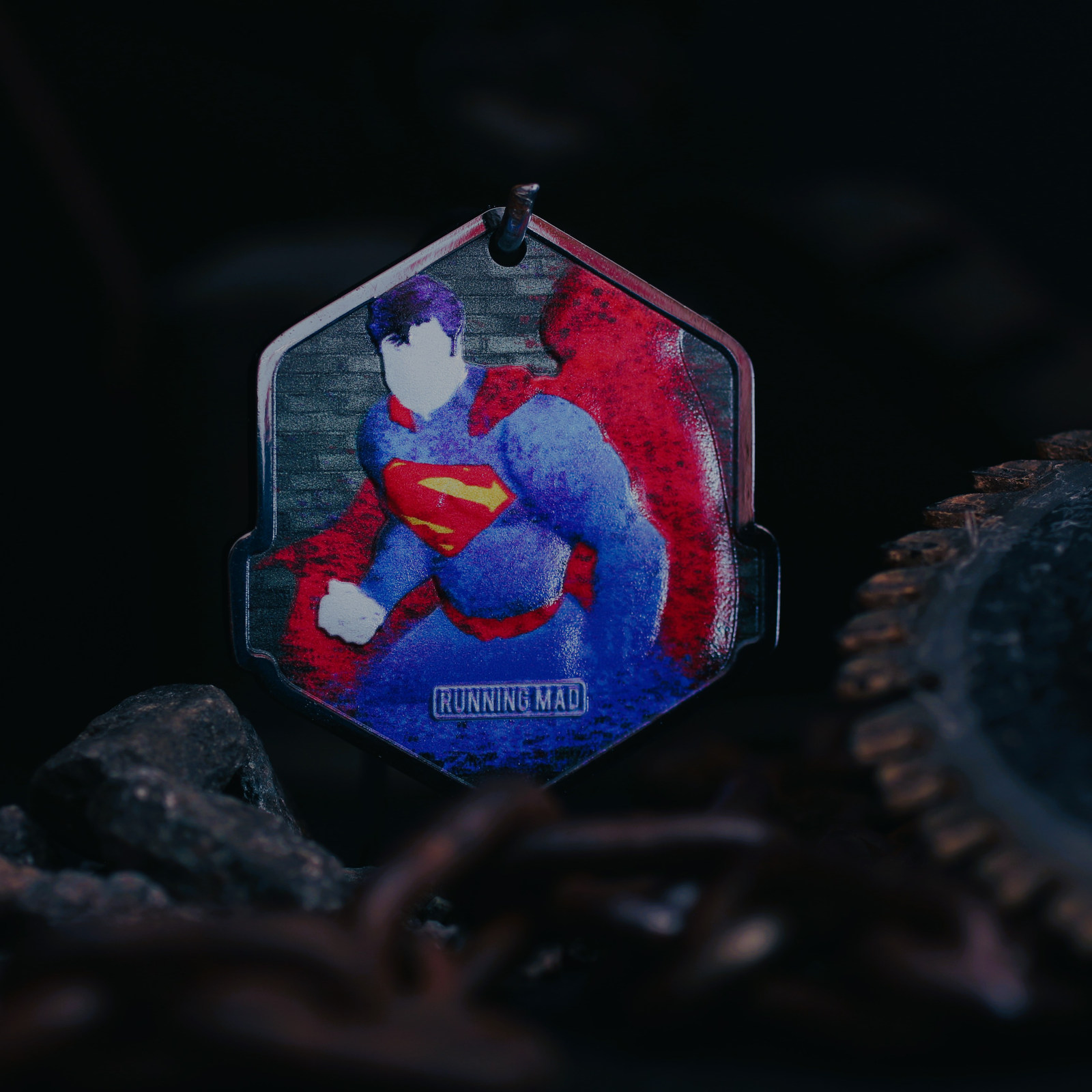 A pendant features a superhero with a red cape and the iconic red and yellow "S" emblem on a blue suit. The background is dark, with a chain and gear parts visible. The text "RUNNING MAD" is at the bottom of the pendant.