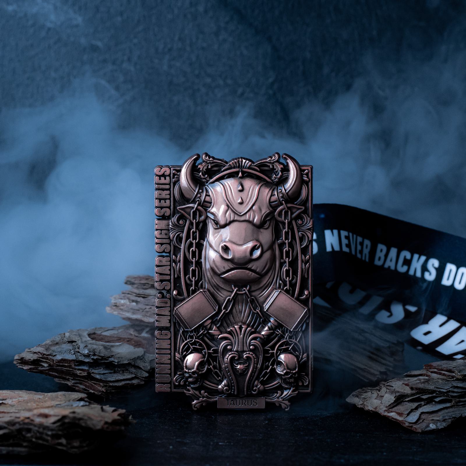 A bronze-colored medallion depicting a bull, surrounded by chains and hammers, standing against a smoky, dark backdrop. The words "TAURUS" and "LIMITED MDL SLSH SERIES" are inscribed. A ribbon with "NEVER BACKS DOWN" in white letters is partially visible.