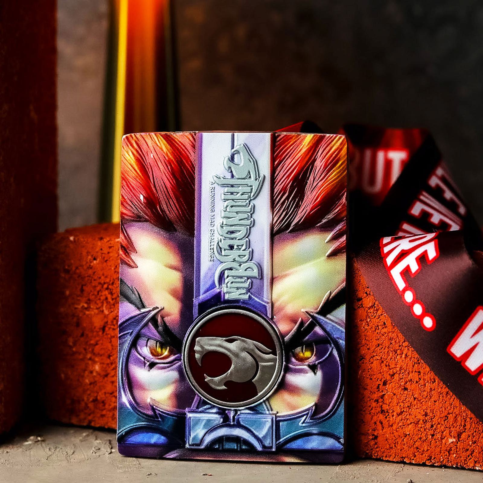 A colorful card featuring a fierce lion face with bright red mane and intense eyes, adorned with a shiny, metallic emblem. The background shows a lit candle, and the card rests between orange bricks. A ribbon is partially visible.