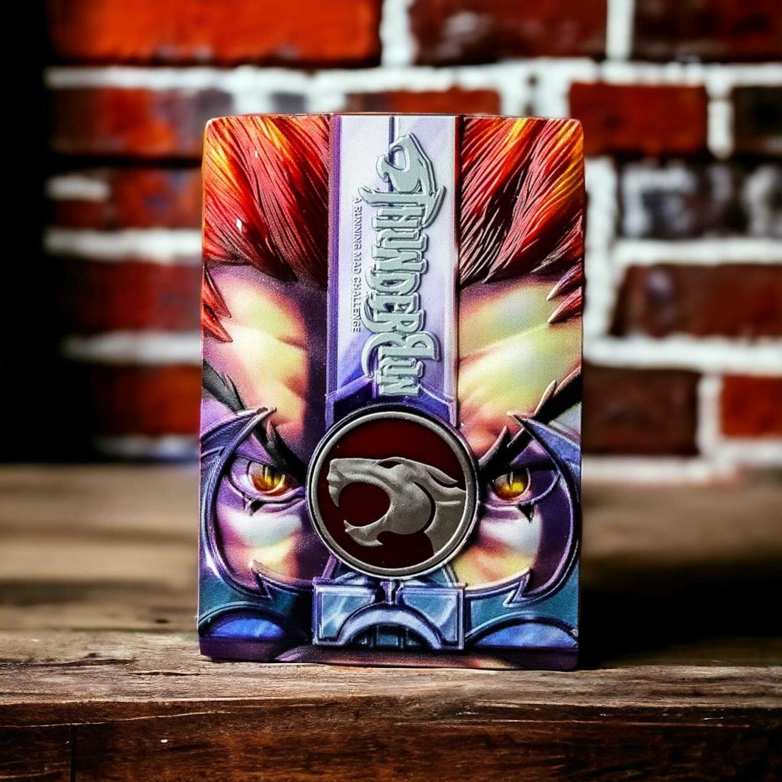A close-up of a colorful box with fierce eyes and a metallic logo featuring a stylized cat head. The box is set against a blurred brick wall background, creating a striking contrast.