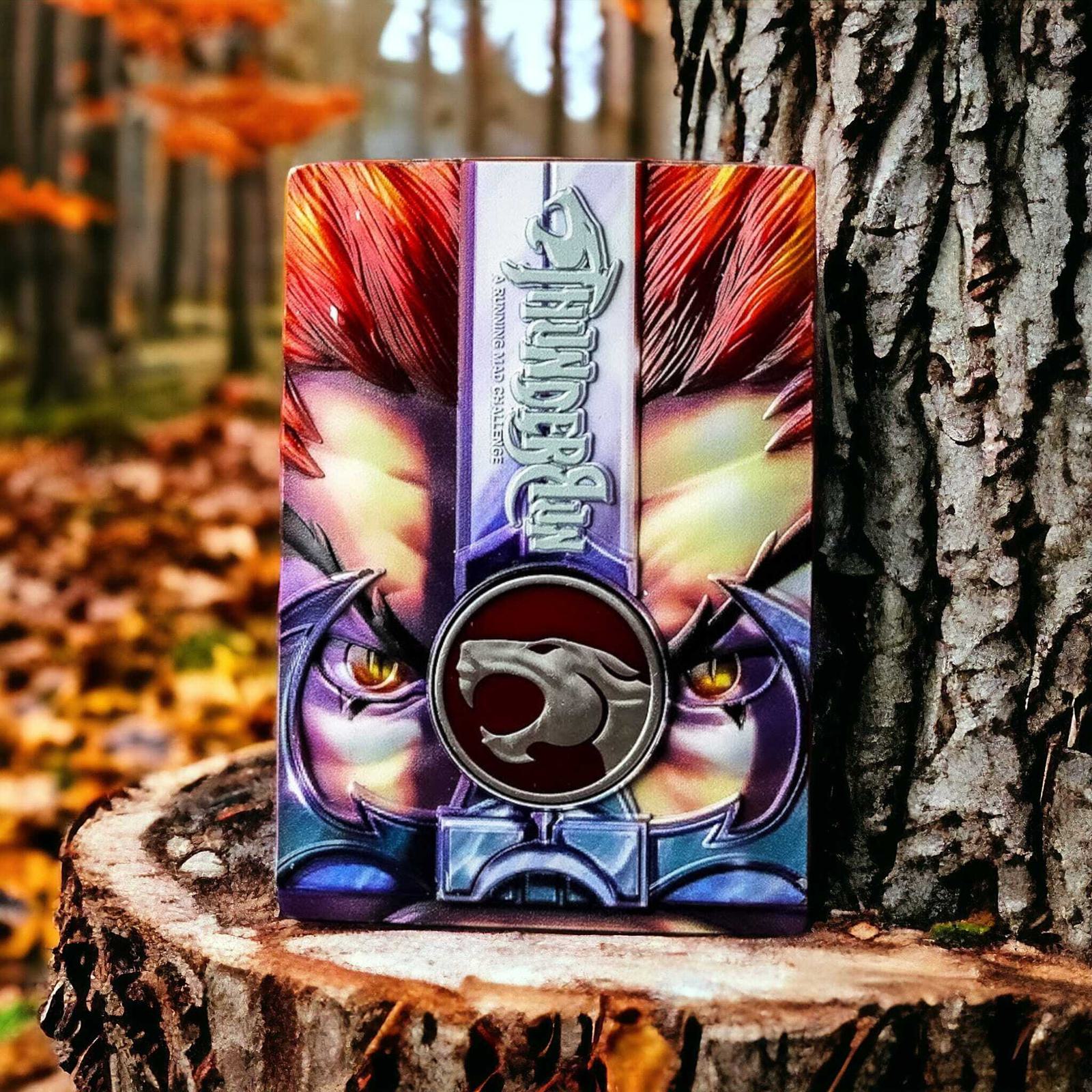 A colorful trading card with intense eyes is propped against a tree trunk in a forest. The card features a fierce lion emblem and vibrant colors, set against a backdrop of autumn leaves on the ground and trees with orange foliage.