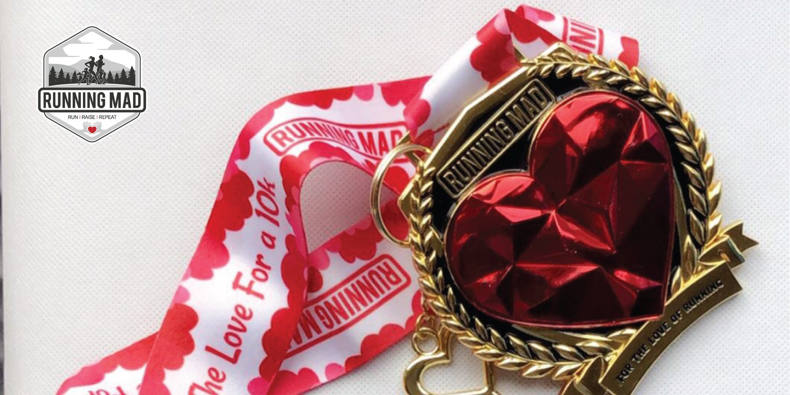 A shiny, heart-shaped medal with a gold trim and a red gem-like center attached to a white and red ribbon. The ribbon features the "Running Mad" logo and the text "Running Mad" and "For the Love of Running." The Running Mad logo is also displayed on the top left.