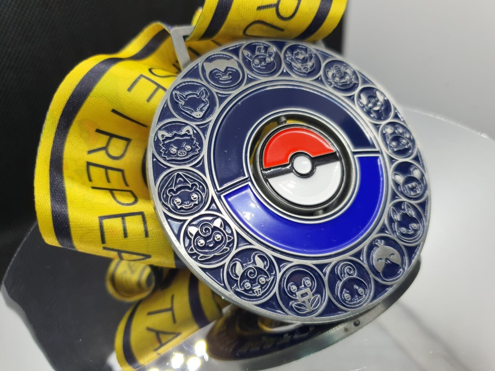 A metallic blue, silver, and red medallion featuring a Poké Ball design in the center encircled by engravings of various Pokémon. The medallion is attached to a yellow ribbon with black text and is placed on a reflective surface.