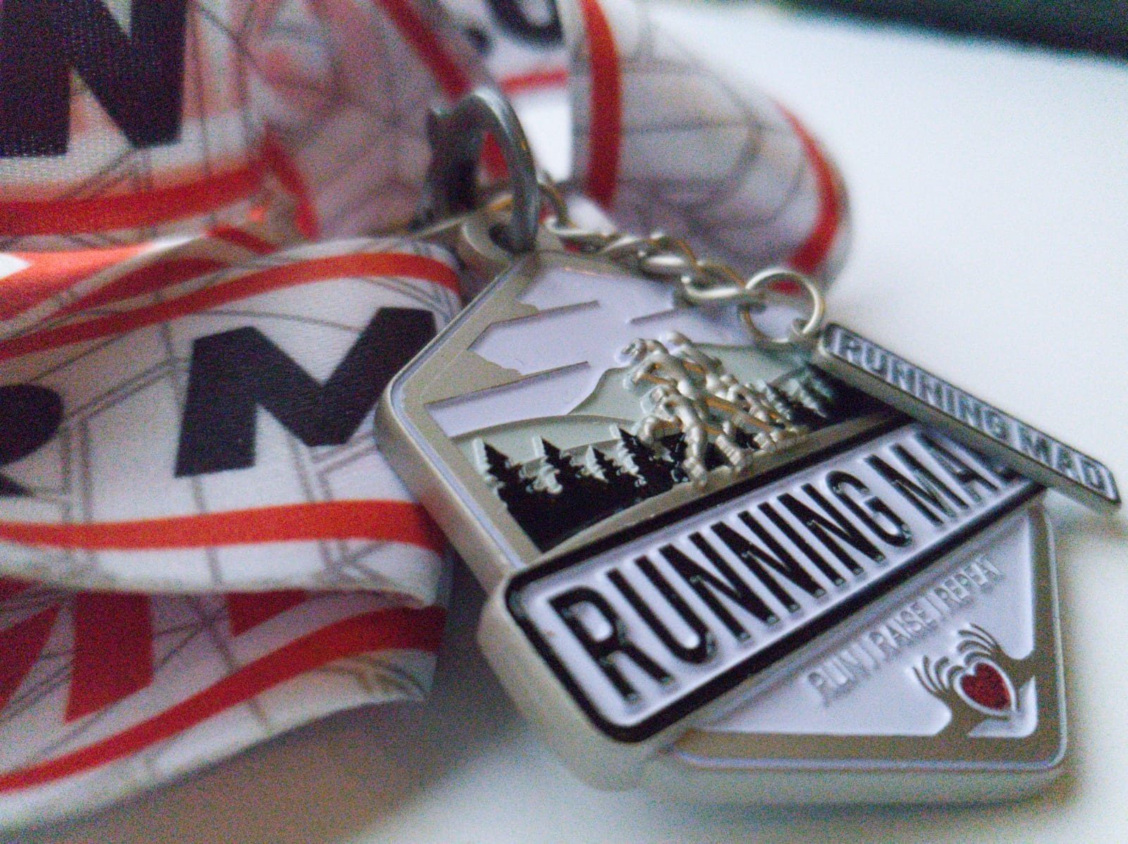 A close-up photo of a silver running medal hanging from a white and red ribbon. The medal features an image of running figures with a forest in the background and the text "RUNNING MAD" along with "RUN FINISH: BEAT" engraved on it.