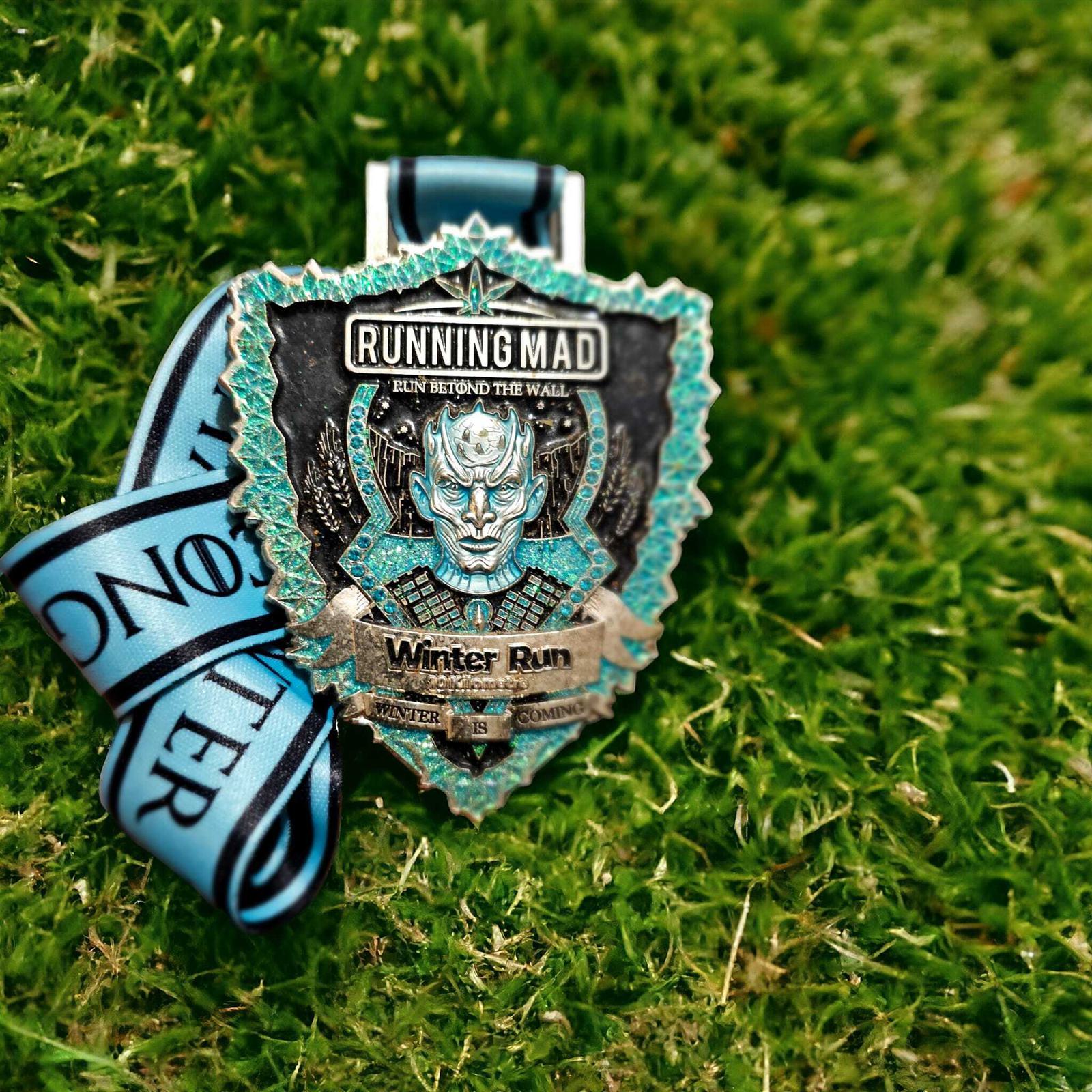 A medal with a blue ribbon lies on green grass. The medal is shaped like a shield and reads "Running Mad Winter Run, Run Beyond the Wall." It features an icy, mythical figure in the center.