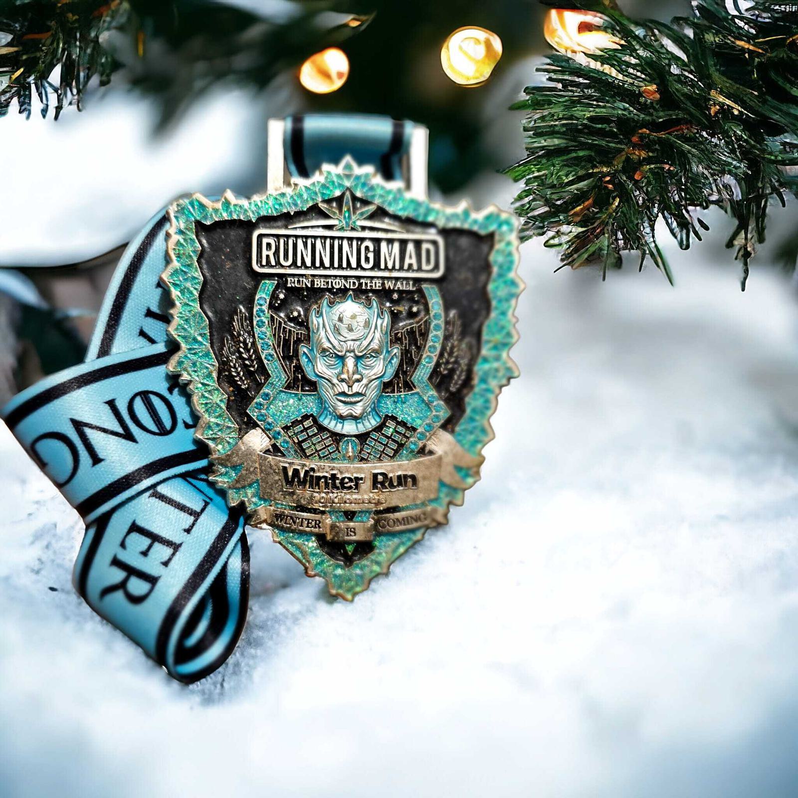 A winter-themed medal hangs on a snow-covered evergreen branch. The medal reads "RUNNING MAD" and "Winter Run" with an icy design. A light blue ribbon displays the word "MONSTER." Warm lights softly illuminate the scene.