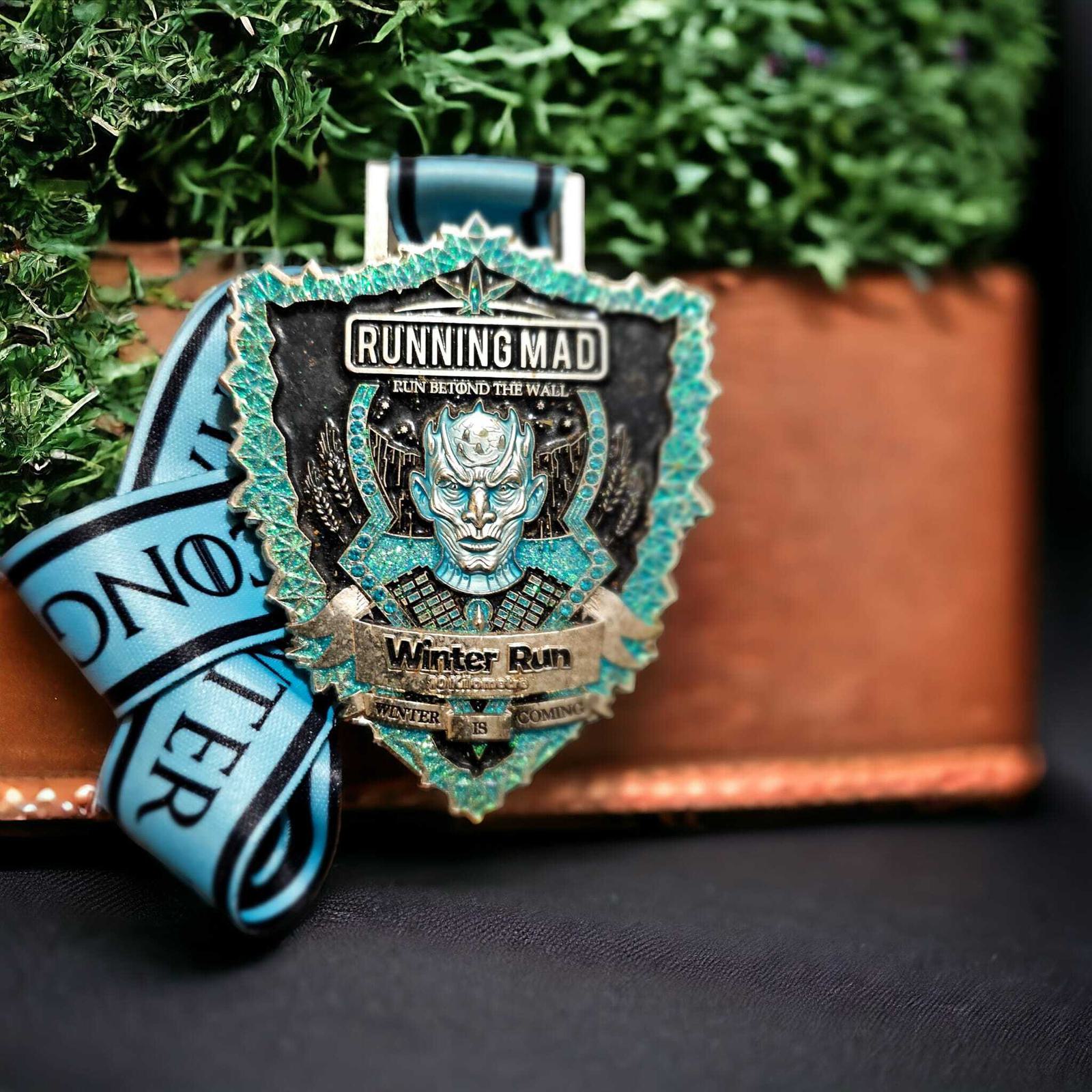 A detailed medal with "Running Mad" and "Winter Run" inscribed, featuring a mythical icy figure. The medal hangs from a blue and black ribbon, set against a green leafy background and a brown base.