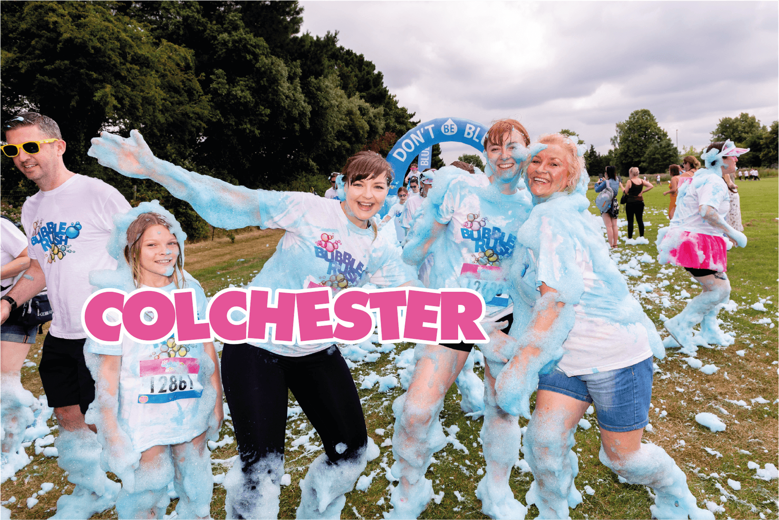 A group of people covered in blue foam are smiling and posing at a fun run event. A young girl stands in front, while three women and one man stand behind her. The text "COLCHESTER" is displayed in bright pink letters. Trees and an inflatable arch are in the background.