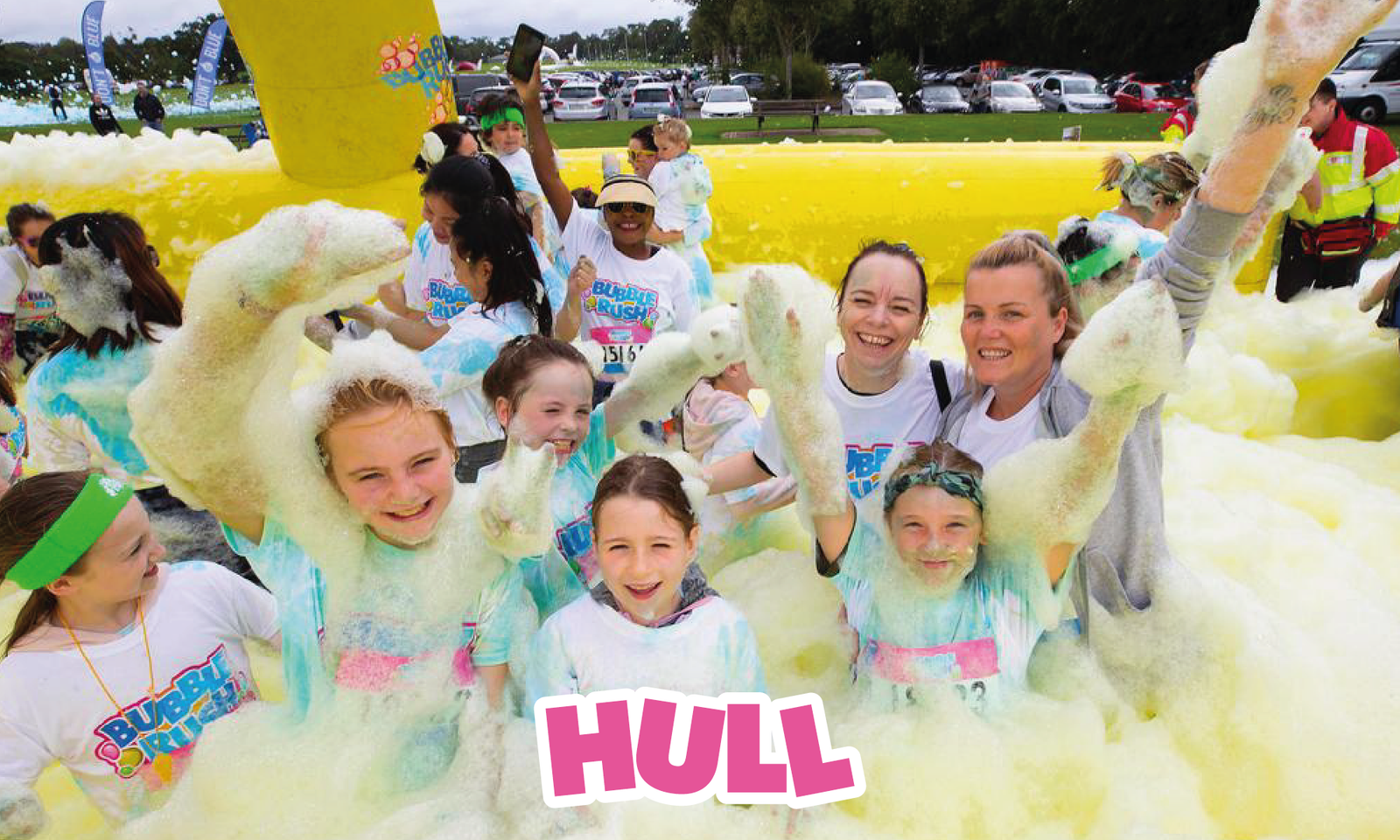 A group of joyful people, mostly children, participate in a foam-filled event wearing white Burst the Rush T-shirts. They are surrounded by yellow foam walls and thick bubbles, with "HULL" in bold pink letters at the bottom center. Everyone is smiling and having fun.