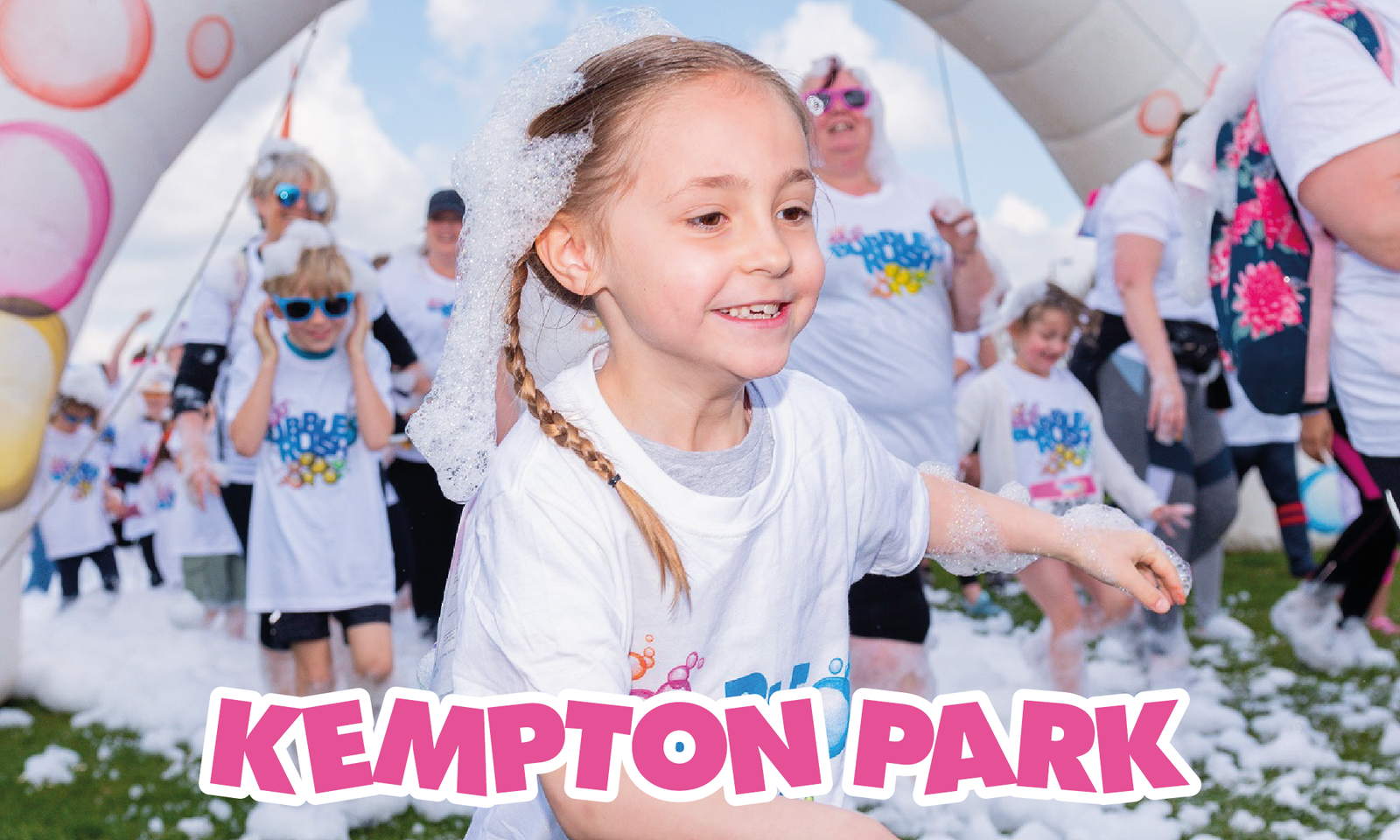 A young girl with braided hair, wearing a white t-shirt, runs amidst foamy bubbles at Kempton Park. Smiling adults and children dressed in similar attire can be seen enjoying the event in the background under a cloudy sky. The text "Kempton Park" is displayed.