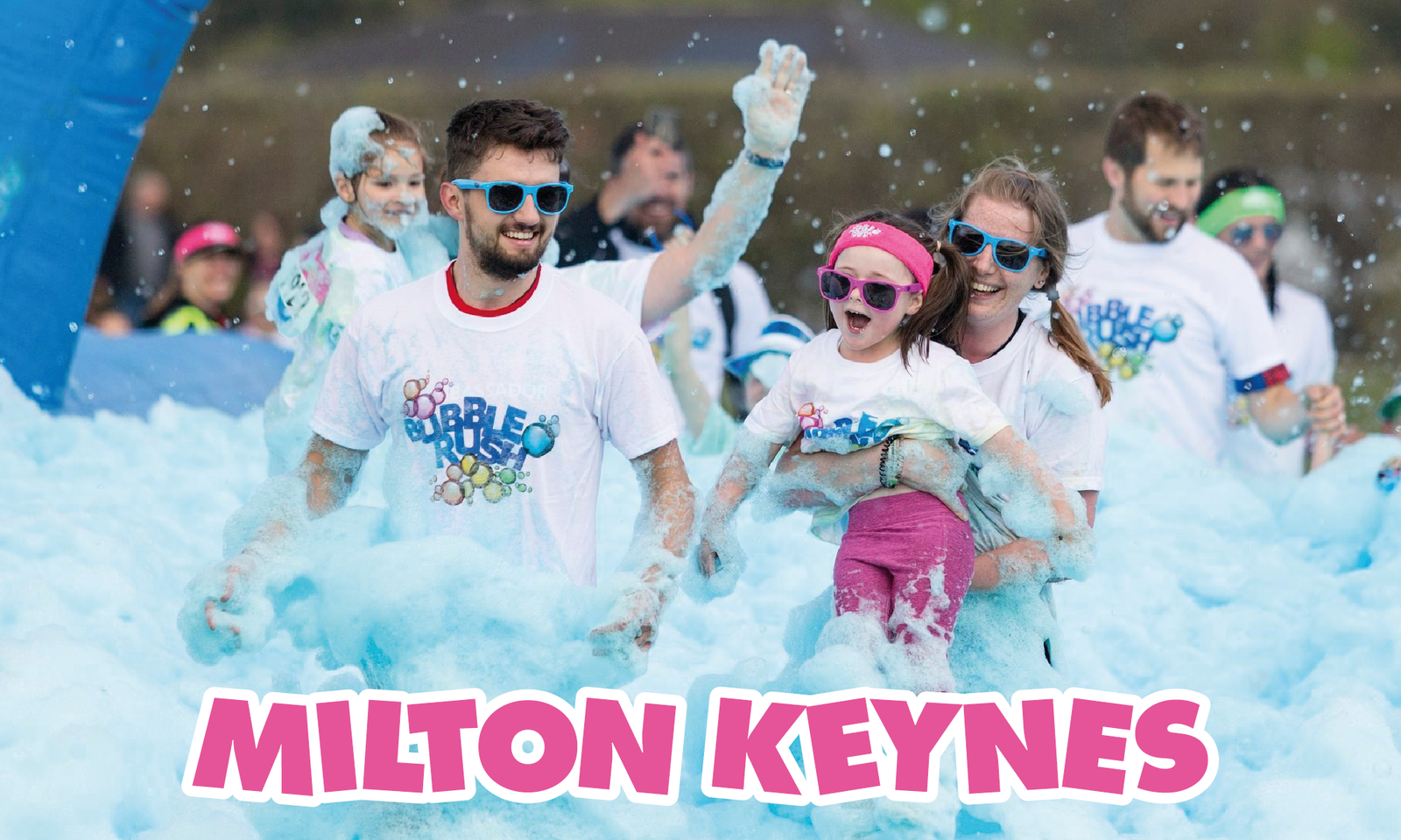 A group of people wearing white t-shirts with "BUBBLES" printed on them and blue sunglasses are joyfully running through colorful foam. "MILTON KEYNES" is boldly displayed at the bottom of the image in pink letters. The mood is festive and playful.
