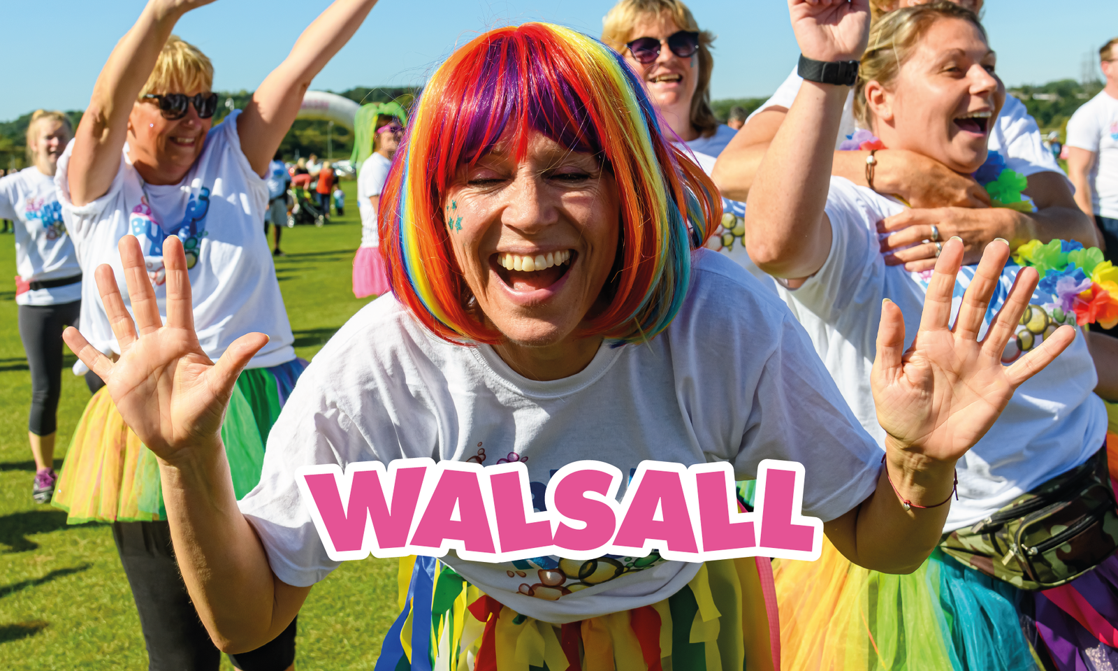 A joyful person in a colorful wig and tutu smiles and raises their hands at an outdoor event. Other participants, also wearing vibrant outfits, laugh and cheer in the background. The word "WALSALL" is prominently displayed across the image.