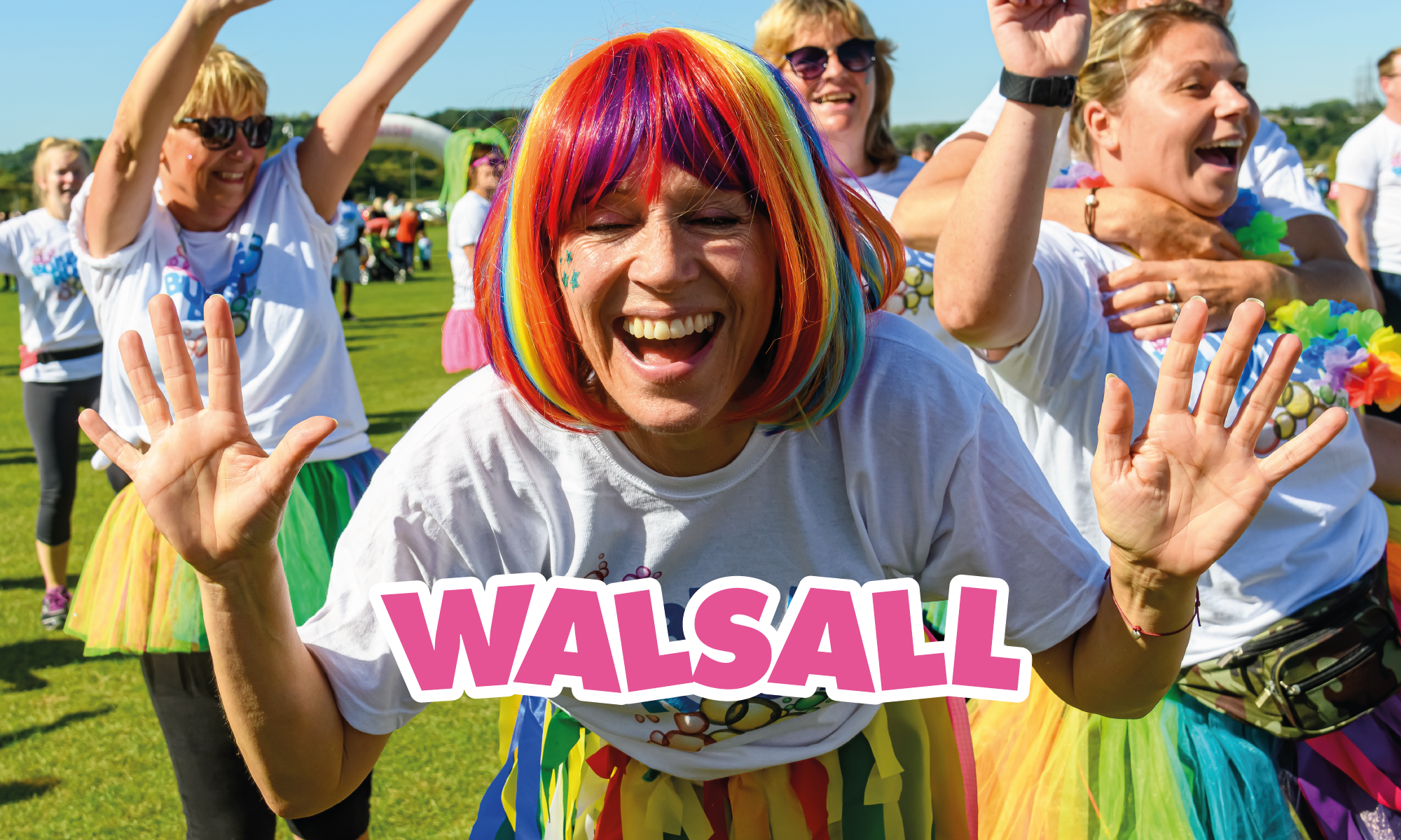 A joyful person in a colorful wig and tutu smiles and raises their hands at an outdoor event. Other participants, also wearing vibrant outfits, laugh and cheer in the background. The word "WALSALL" is prominently displayed across the image.