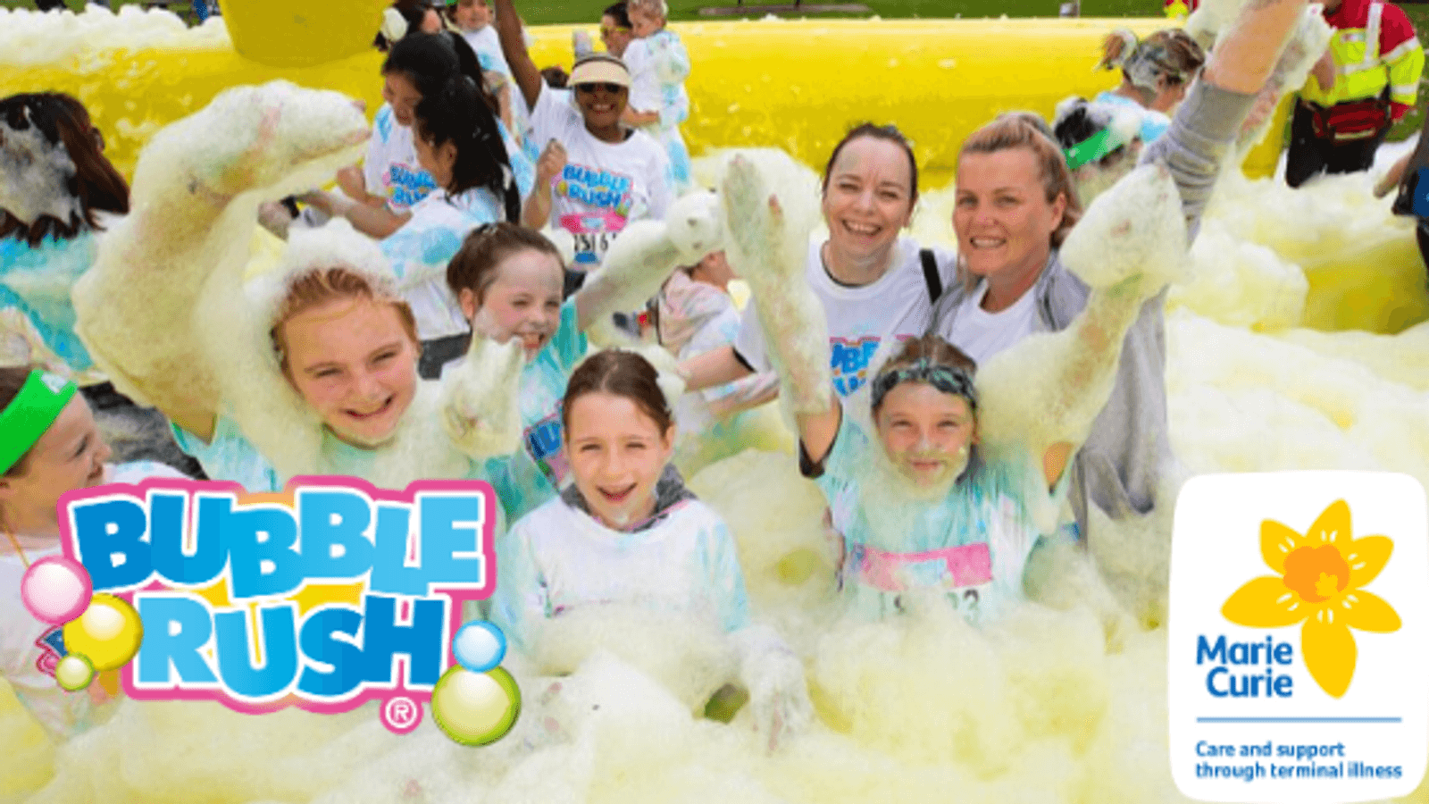 A cheerful group of people, including children and adults, playfully enjoying a foamy environment during a Bubble Rush event. They are surrounded by white and yellow foam, raising their arms and smiling at the camera. The Bubble Rush and Marie Curie logos are displayed in the image.