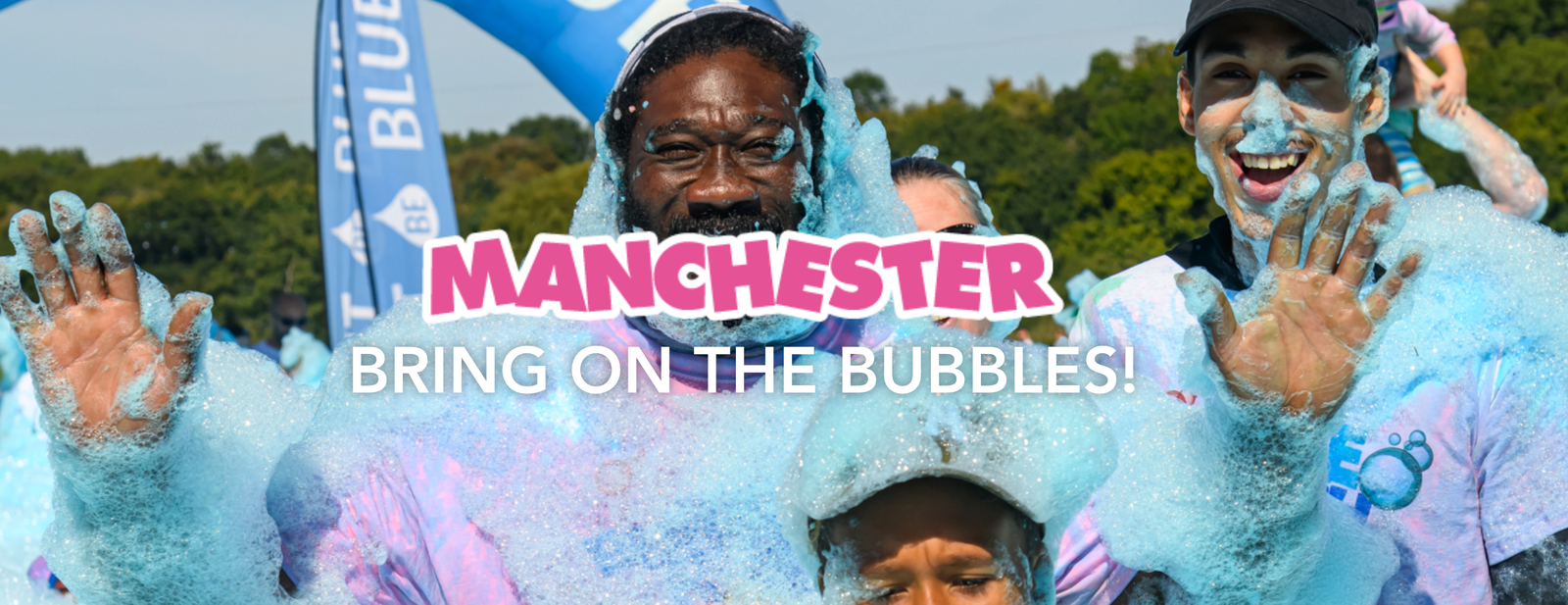 Participants at a bubble-themed event in Manchester, covered in blue foam, smile and wave at the camera. The background shows trees and event banners. Text overlays read "MANCHESTER" and "BRING ON THE BUBBLES!.