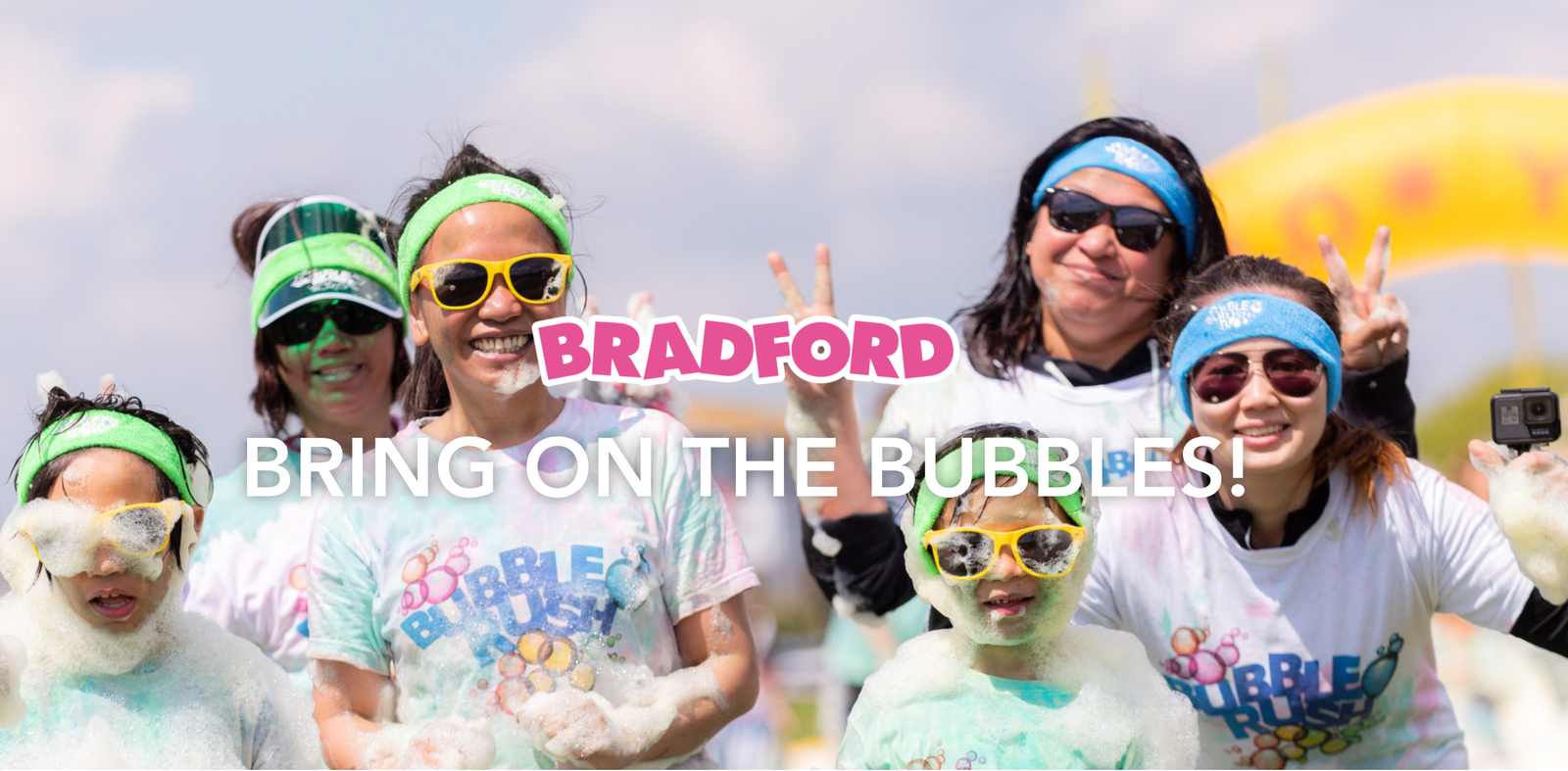 A group of people wearing white 'Bubble Rush' t-shirts and colorful sunglasses smile and pose with peace signs, surrounded by soap bubbles. Text on the image reads "BRADFORD" and "BRING ON THE BUBBLES!" in bold letters.