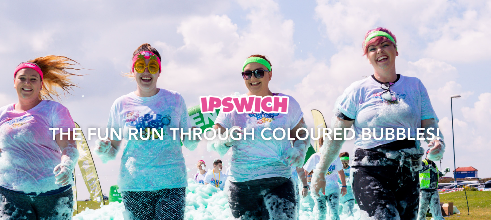Four people, wearing headbands and colorful sunglasses, run through a plethora of colored bubbles during the Ipswich Bubble Rush fun run event. They are smiling and laughing under a bright blue sky dotted with fluffy clouds. Signage in the background reads, "THE FUN RUN THROUGH COLOURED BUBBLES!.