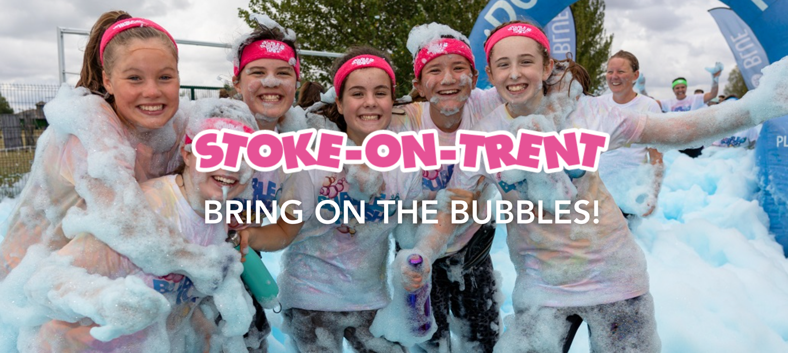 A group of smiling women and girls, covered in foam, wearing pink headbands and white shirts, stand together outdoors at a foam run event. The text reads, "STOKE-ON-TRENT BRING ON THE BUBBLES!" A blue banner and trees are visible in the background.