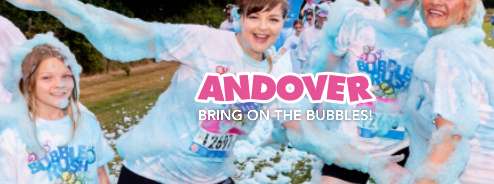 Participants in white 'Bubble Rush' T-shirts run through blue foam outdoors, smiling and cheerful. The text "ANDOVER BRING ON THE BUBBLES!" is displayed prominently. The event appears fun and festive, with many people engaging in the activity.