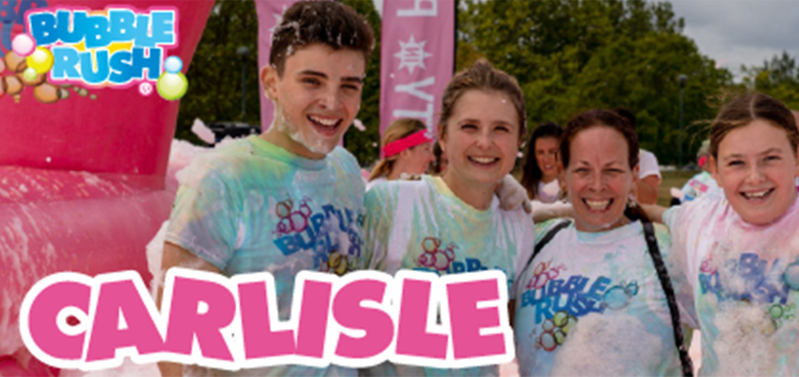 A group of four people, two adults and two children, are smiling and standing together covered in colorful foam at a Bubble Rush event in Carlisle. They are wearing white Bubble Rush t-shirts, and there are trees and event signs in the background.