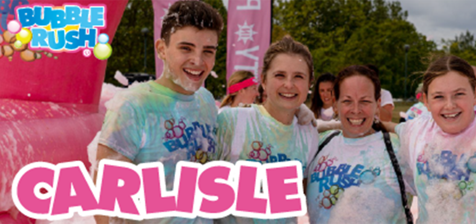 A group of four people, two adults and two children, are smiling and standing together covered in colorful foam at a Bubble Rush event in Carlisle. They are wearing white Bubble Rush t-shirts, and there are trees and event signs in the background.