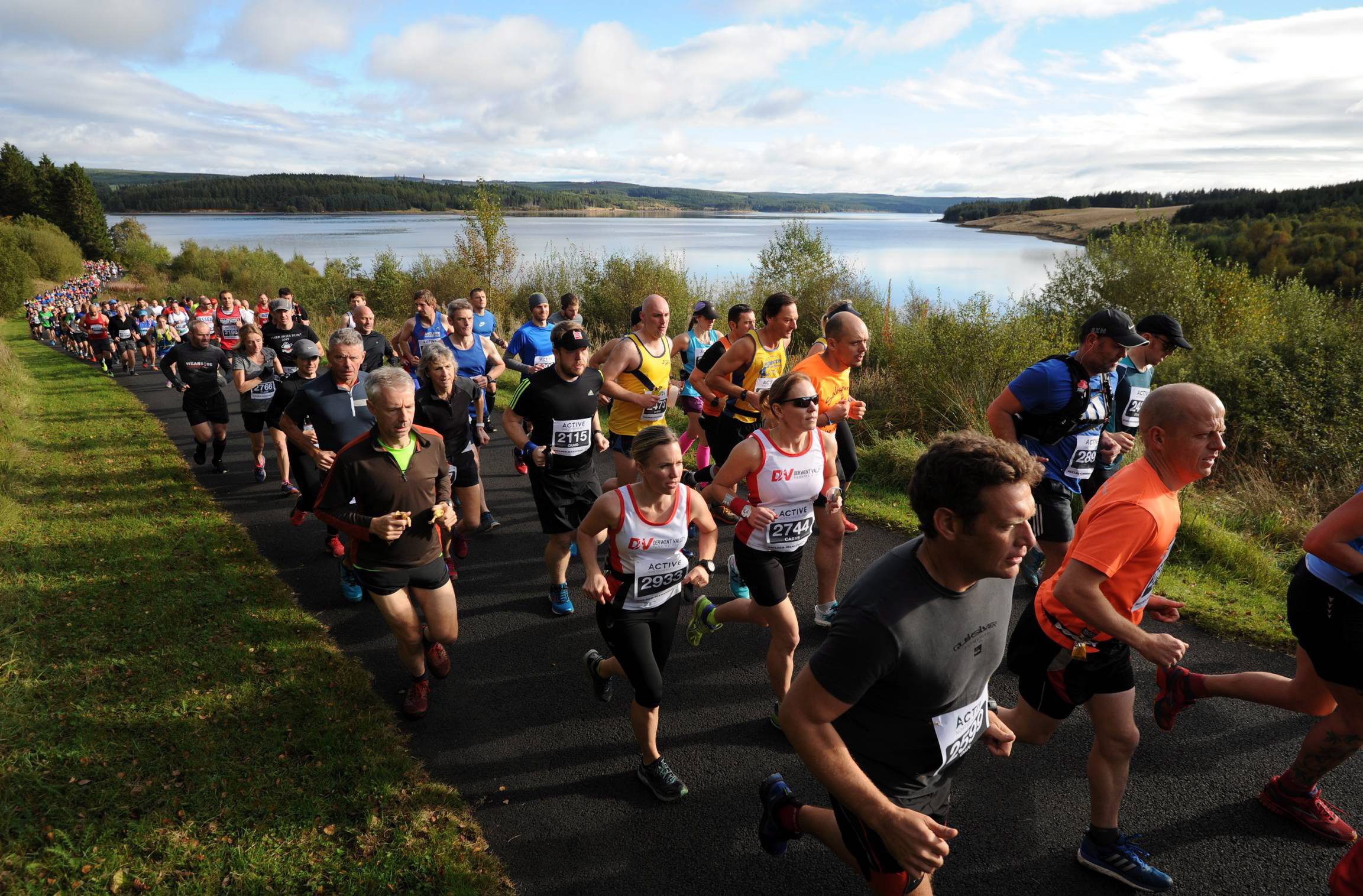 A large group of runners participate in a marathon on a paved road along the edge of a scenic lake, surrounded by lush greenery and under a partly cloudy sky. The participants wear various athletic attire and appear focused on their run.
