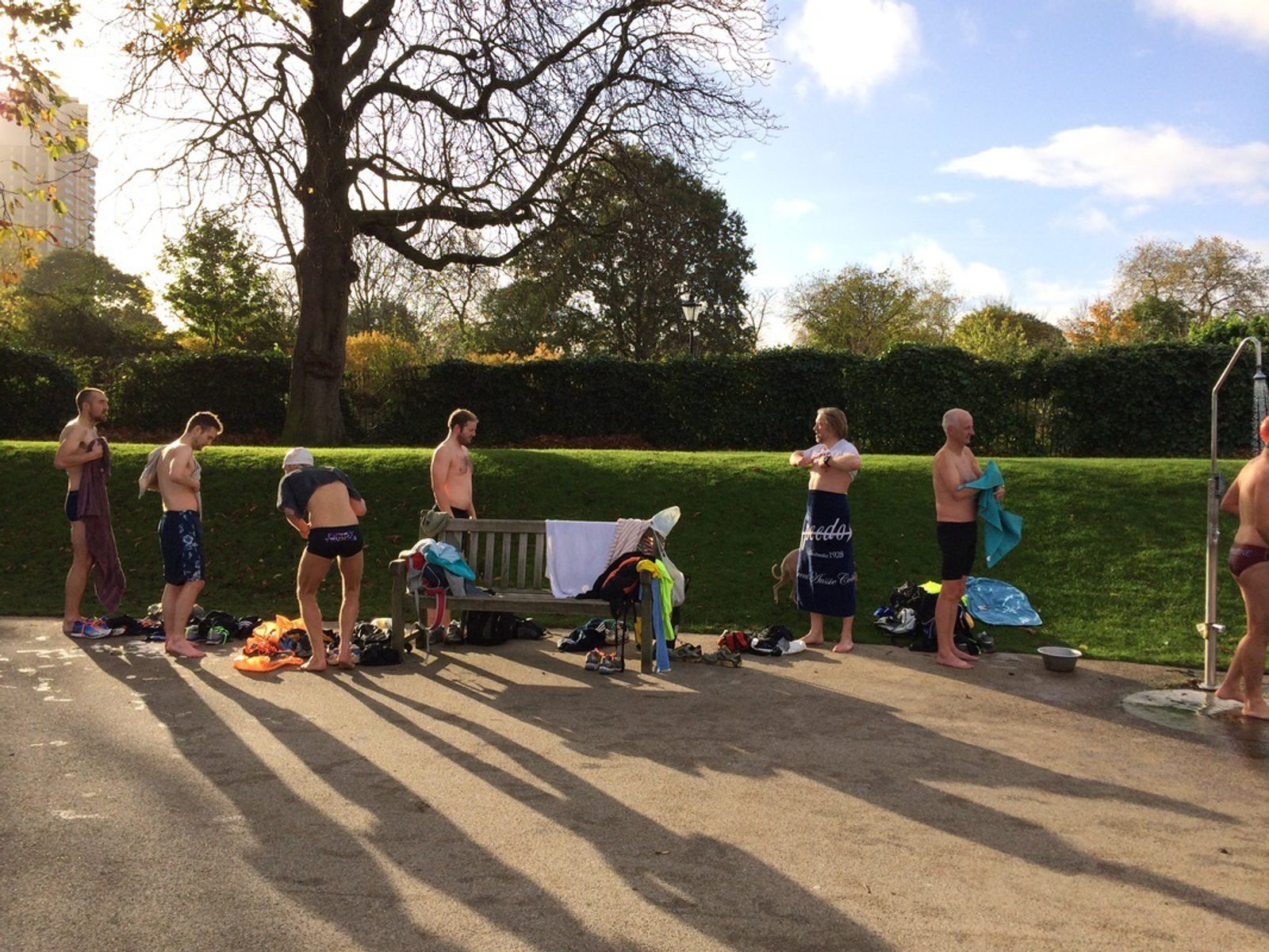 A group of people stand in a line outdoors, changing into their swimwear near a park bench. Some are already in swimsuits, while others are partially dressed. The weather is sunny, and there are trees and a grassy area in the background.