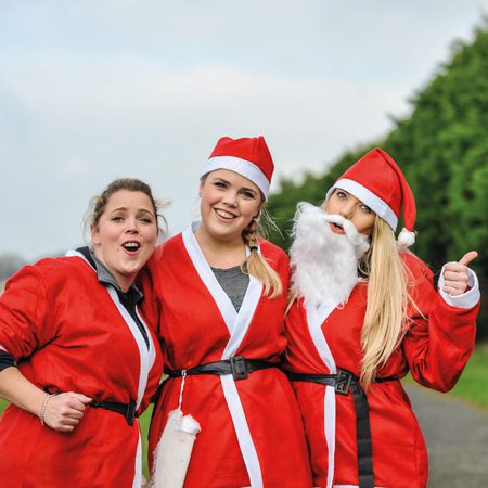 Three people are standing outdoors, dressed in red Santa costumes with white trim. One person is wearing a Santa hat and beard, while the others are smiling and posing, with one giving a thumbs-up. They appear to be enjoying a festive occasion. Trees are in the background.