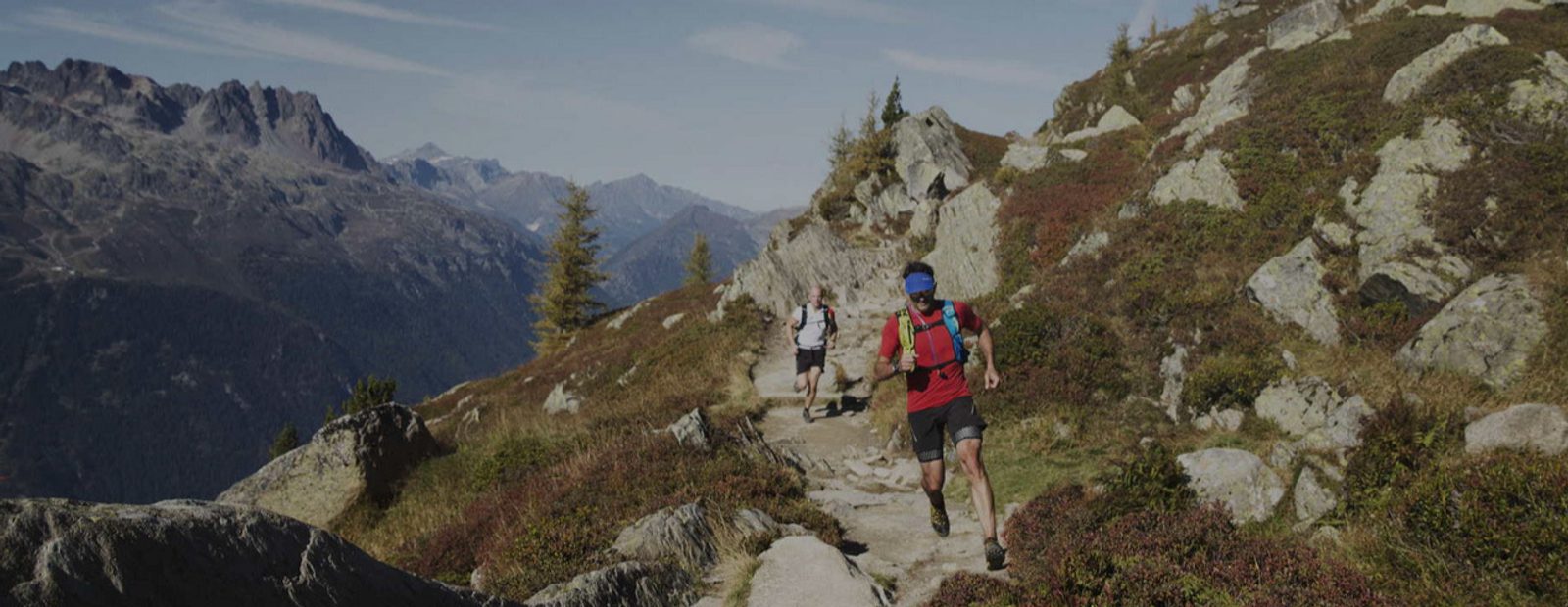 Two trail runners traverse a rocky mountain path surrounded by rugged terrain and high peaks. The sky is clear blue, and the landscape features sparse vegetation and jagged rocks. The lead runner wears a red shirt, and the follower wears a white shirt.