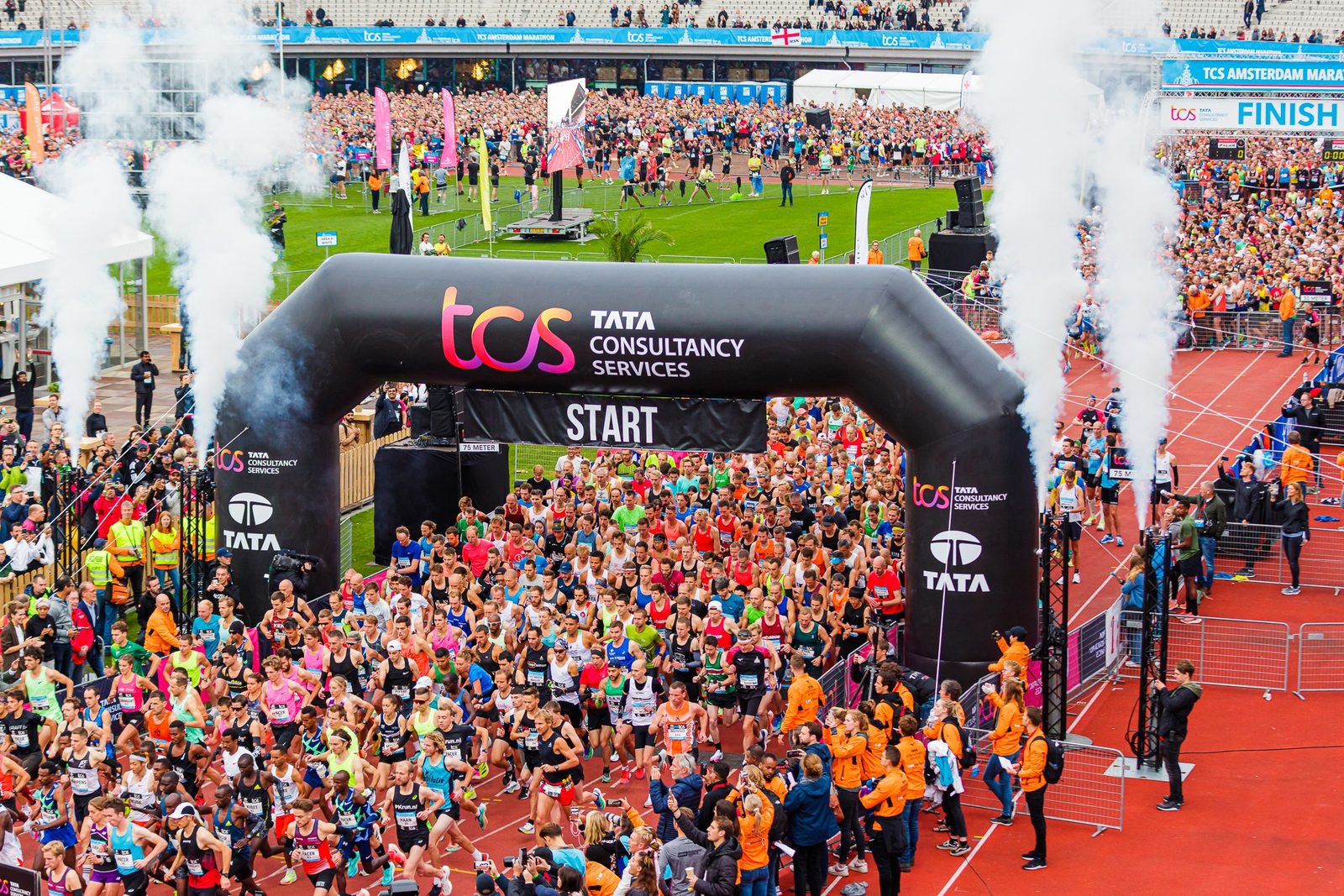 A large crowd of runners begins a race under a black and pink "Tata Consultancy Services" start banner on a track, with smoke flanking the sides. Spectators and officials wearing orange jackets line the edges. In the background, the finish line is visible.