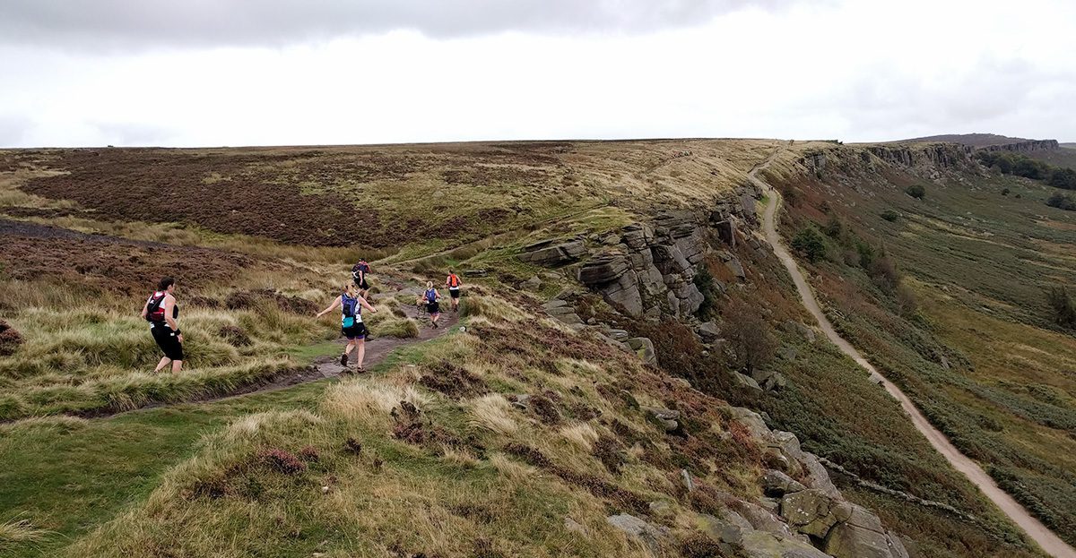 Runners on a narrow, winding trail along the edge of a rocky cliff in a hilly, grass-covered landscape. The sky is overcast, and the terrain varies between grassy patches and rough, rocky sections. Some runners are closer while others are farther ahead on the trail.