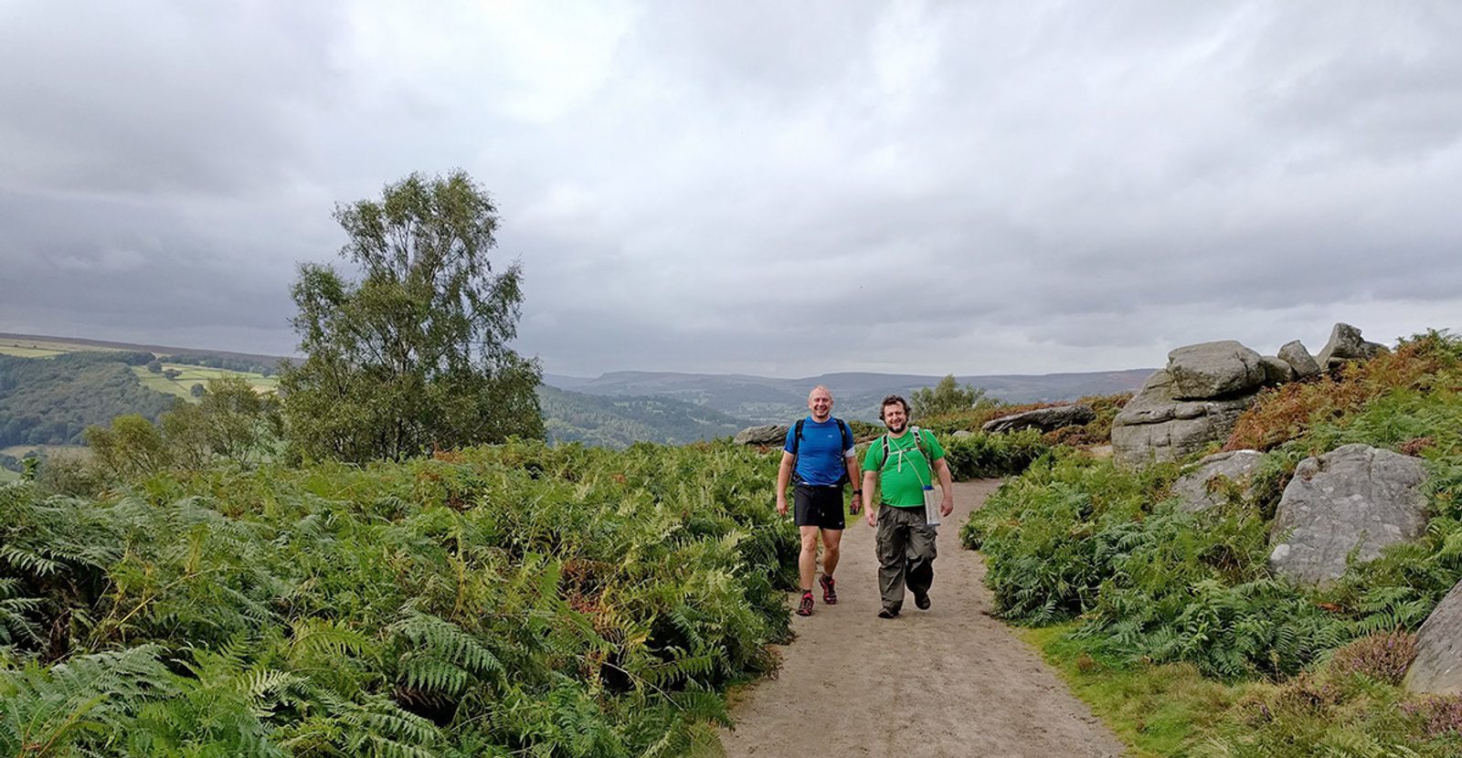 Two men in hiking attire walk along a dirt path surrounded by lush green ferns and large rocks. A scenic, hilly landscape stretches out behind them under a cloudy sky.