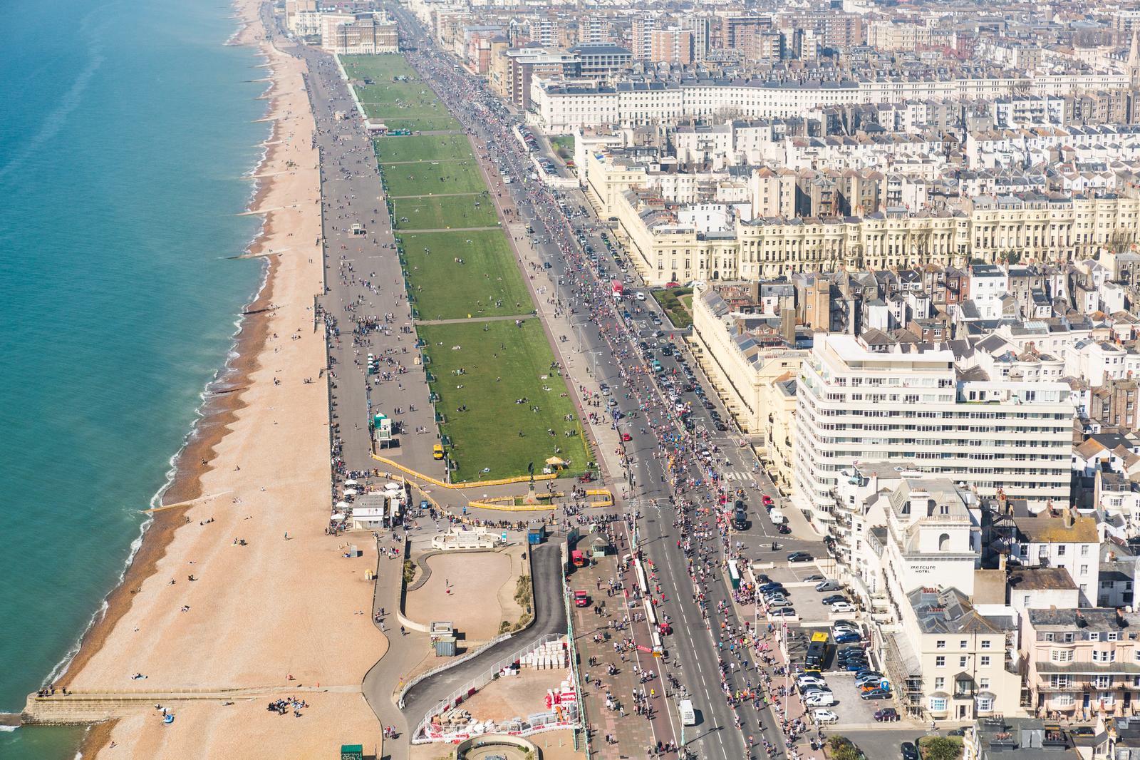 Aerial view of a coastal city with a beach on the left and an expansive urban area on the right. A promenade and road separate the beach from the city, with numerous people walking, cycling, and resting along the beach and greenery. Buildings and sidewalks are busy.